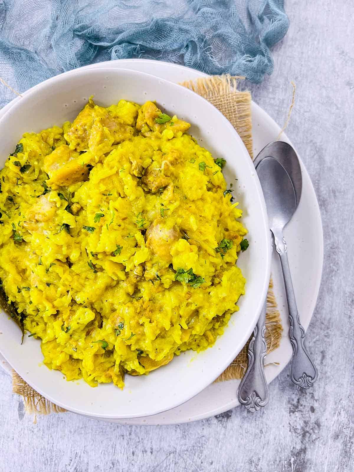 Bowl of chicken khichdi on a white plate with spoons in the background.