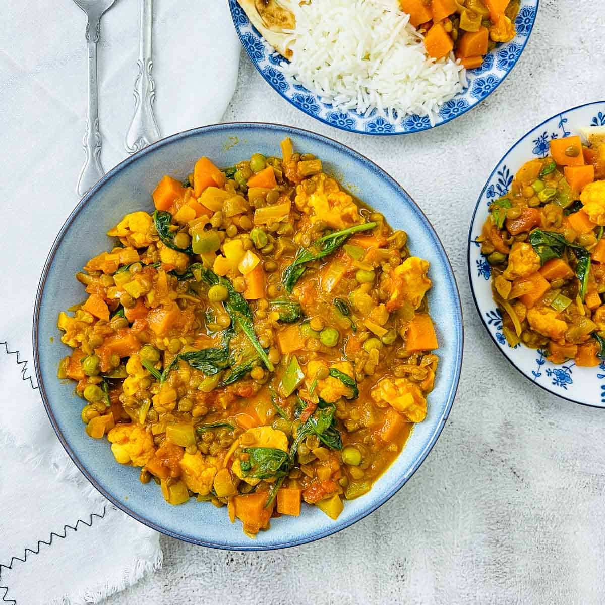 Vegetable lentil curry in a blue bowl with two plates of rice and curry in the background.