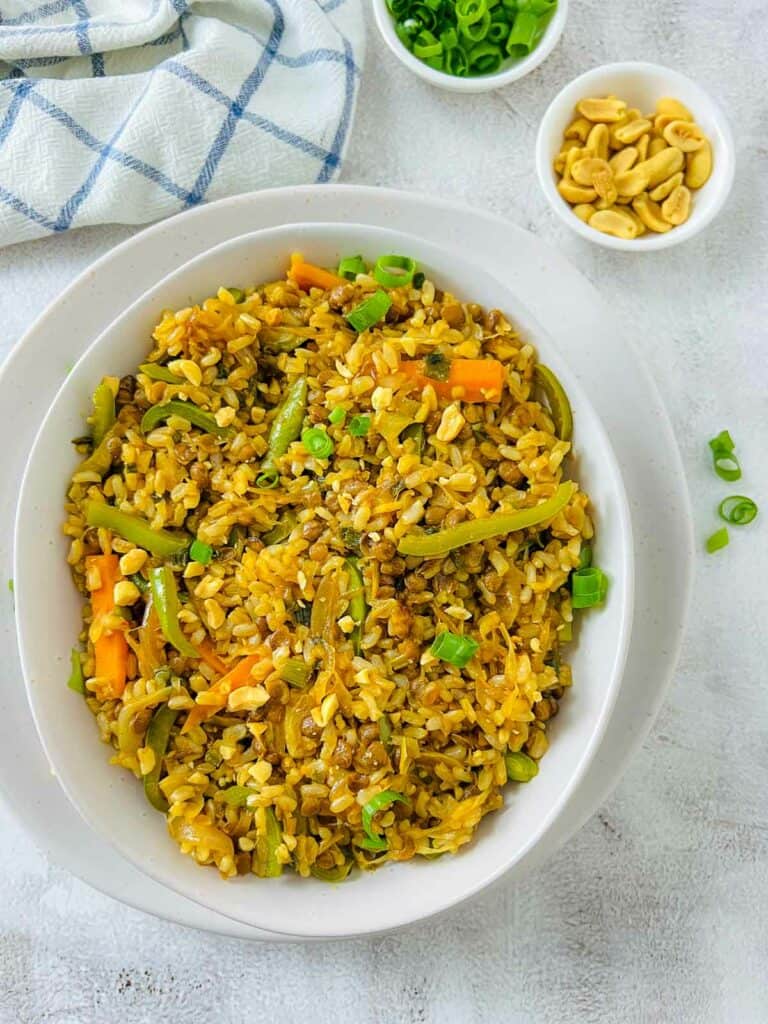 Brown rice and lentils stir fry in a white bowl with roasted peanuts and scallions in the background.