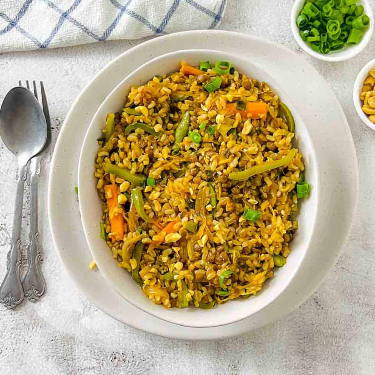 Brown rice and lentils stir fry in a white bowl with spoons and scallions on the side.