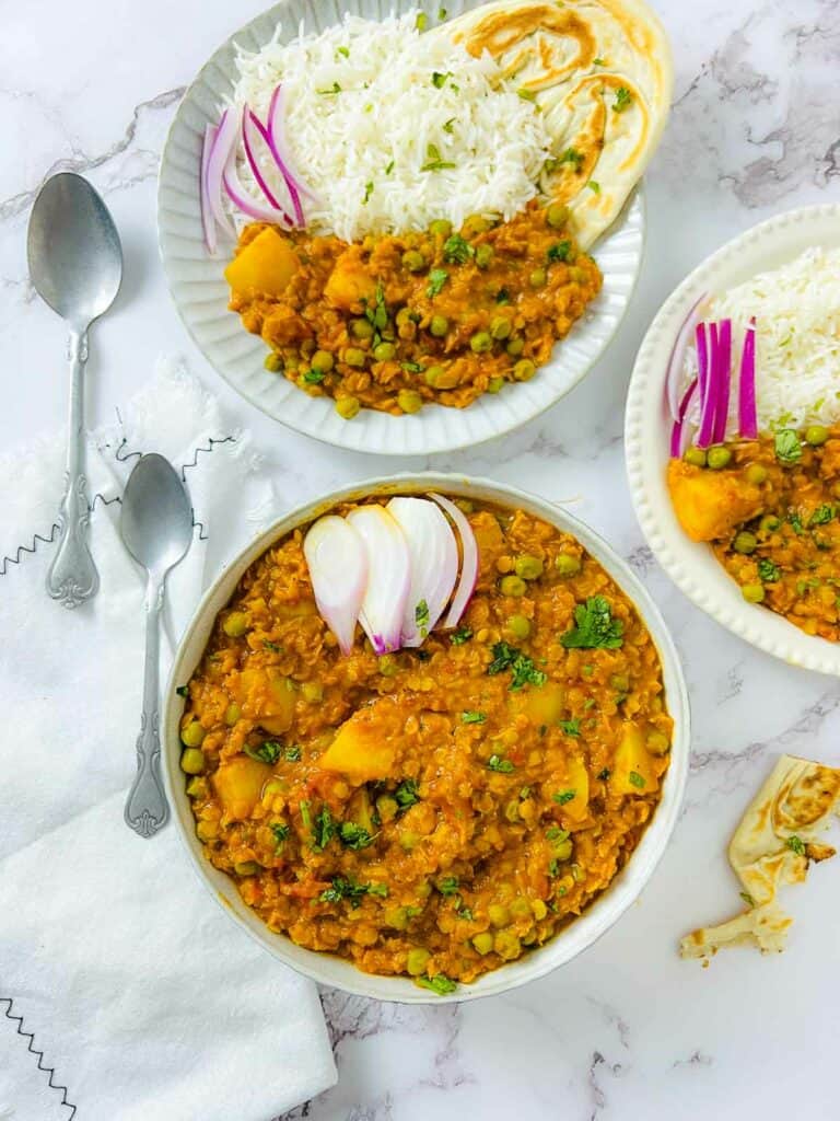 Lentil peas potato curry in a white bowl with two plates with rice and naan in the background.