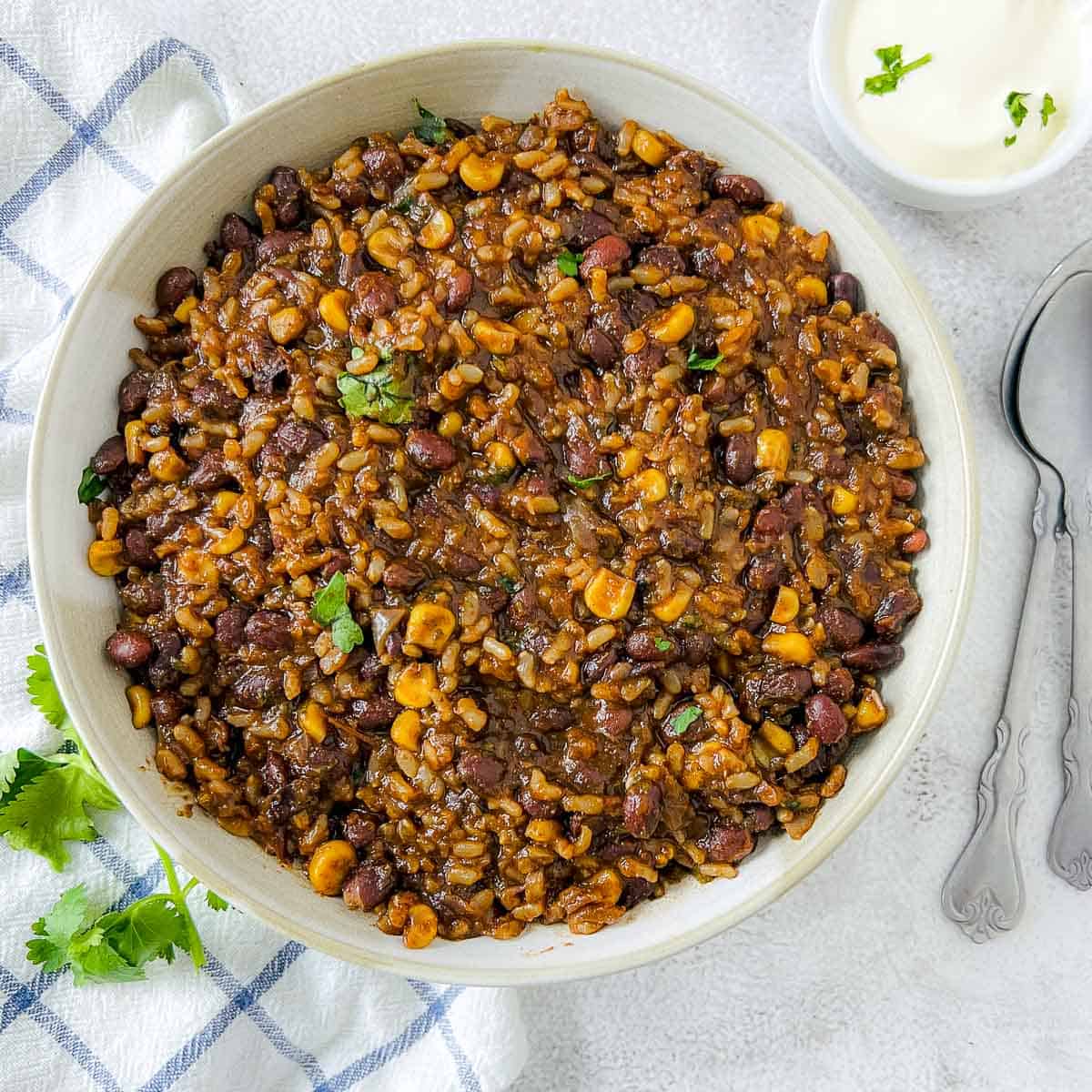 Mexican rice and beans in a white bowl with sour cream and spoons in the background.
