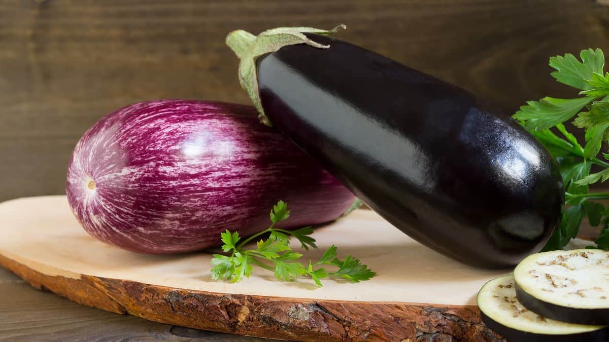 Eggplants placed on a wooden board with herbs in the background.