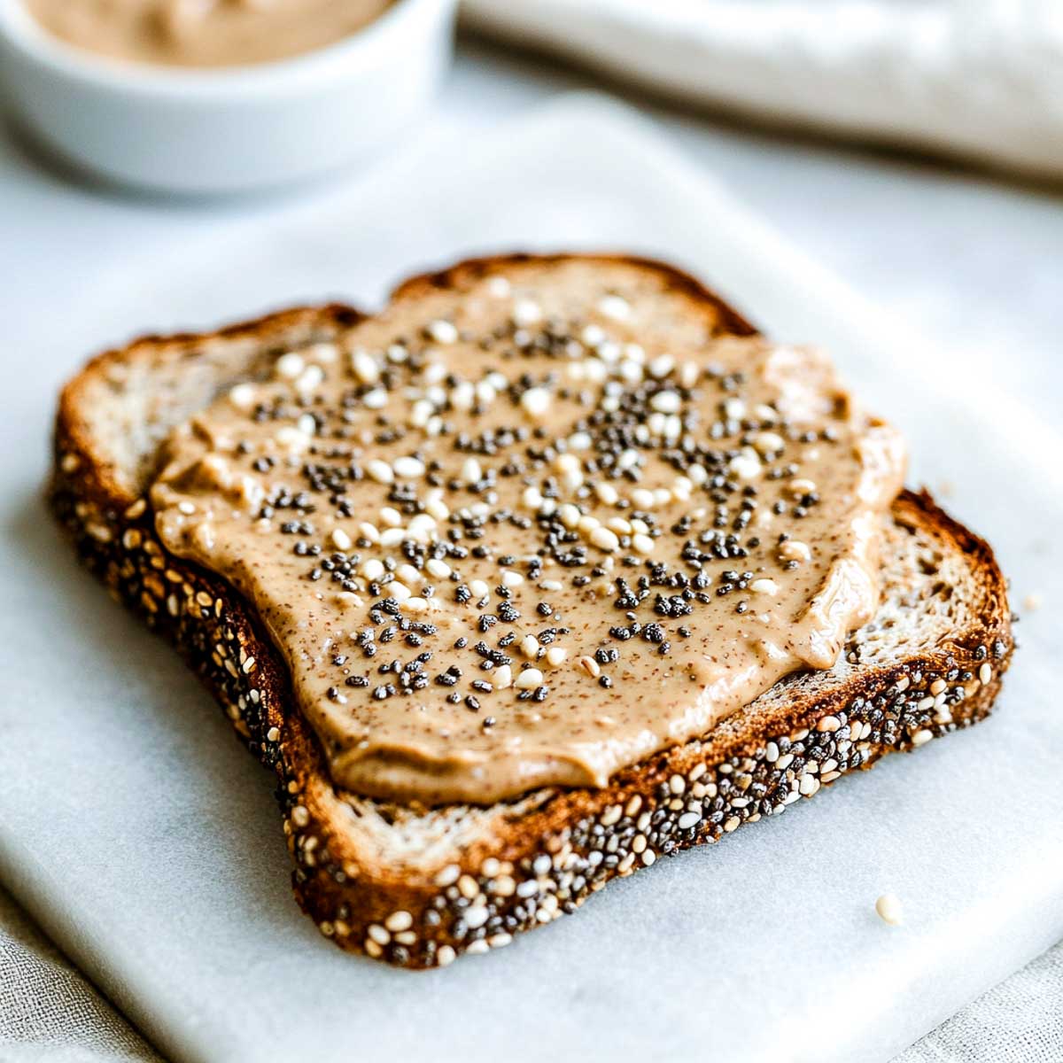 A slice of whole grain toast with creamy almond butter, topped with chia seeds, hemp hearts, and a drizzle of honey, placed on a white marble surface.
