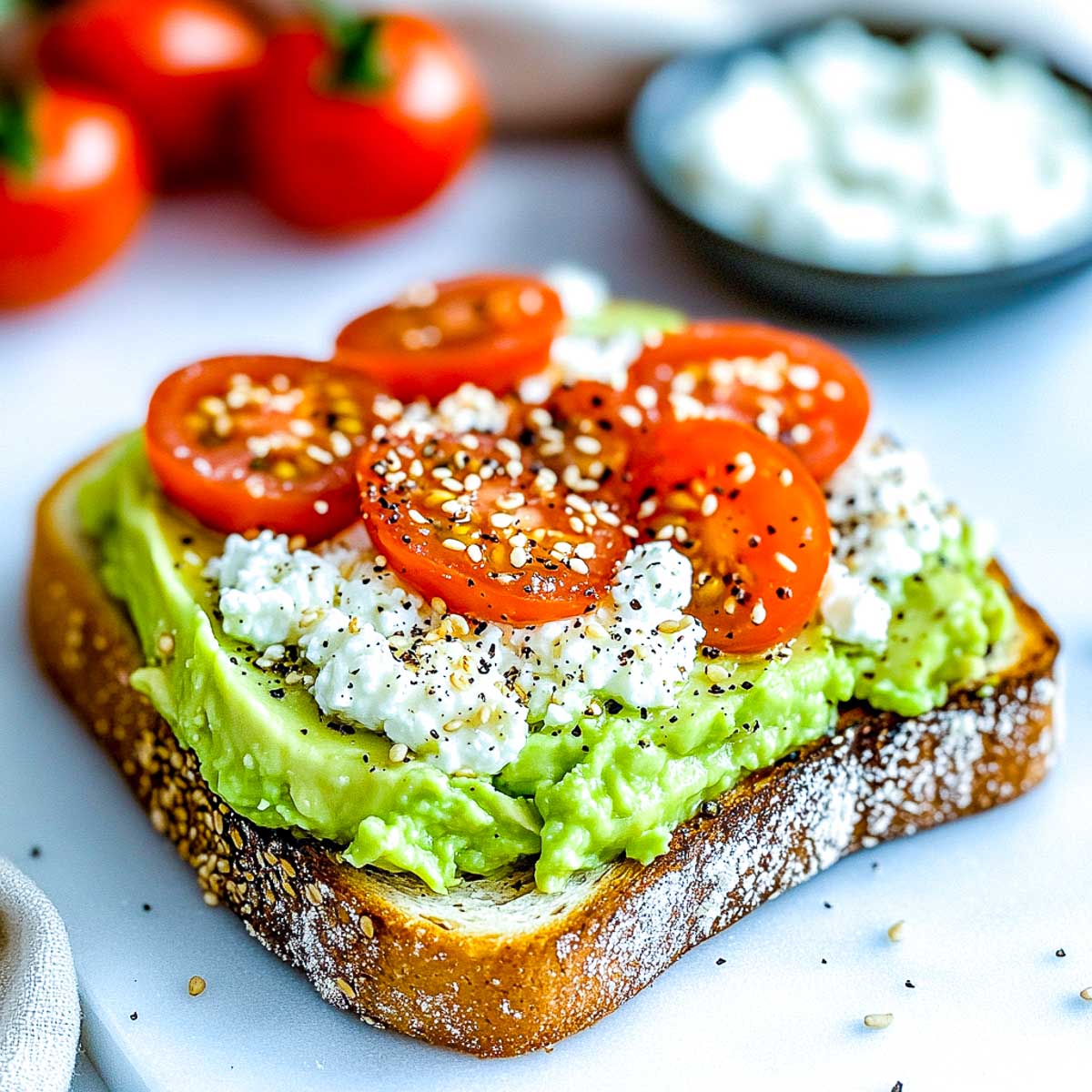 A slice of sourdough toast topped with mashed avocado, cottage cheese, cherry tomatoes, and sesame seeds, placed on a white marble surface.
