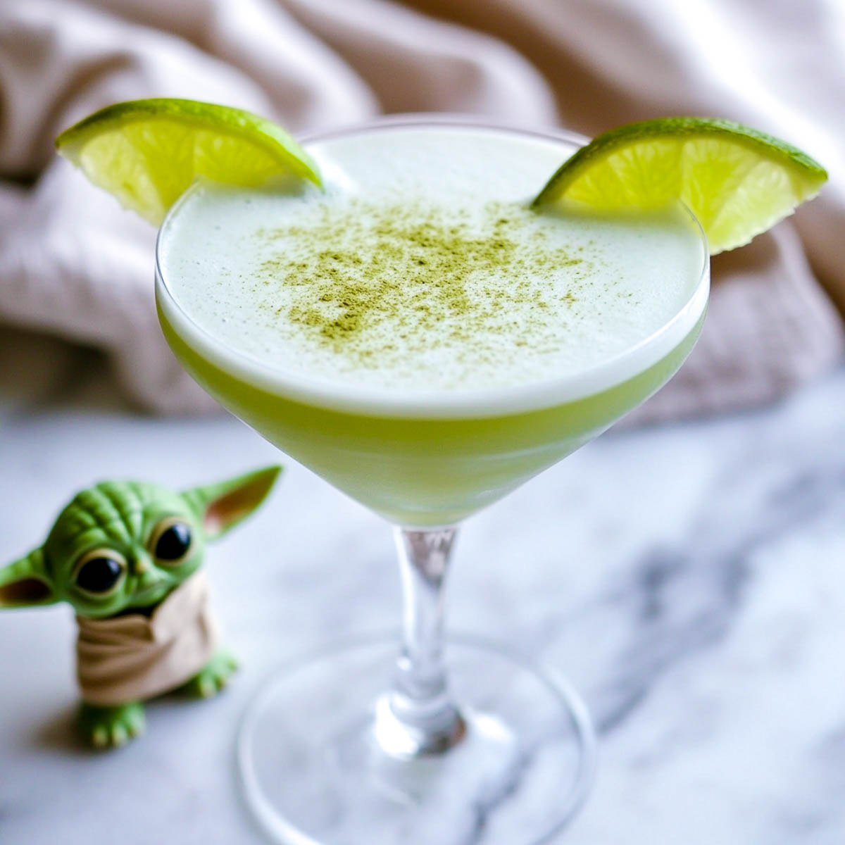 A pale green Baby Yoda-inspired cocktail in a martini glass, garnished with two lime wedges resembling ears, and a light dusting of matcha powder. The drink is placed on a white marble countertop, with a beige linen napkin in the background.