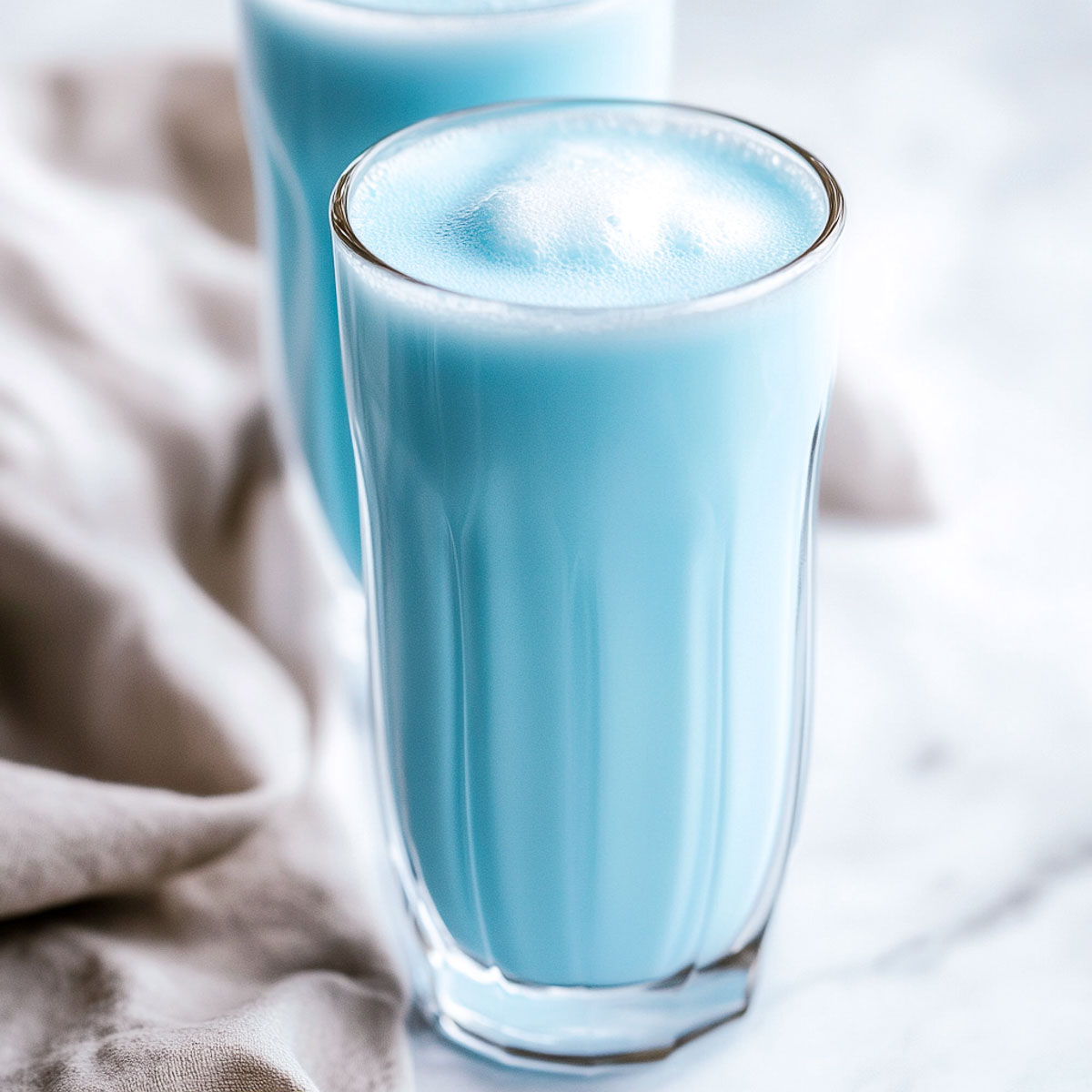A chilled glass of creamy blue milk served in a tall clear glass on a white marble surface. The drink has a smooth, pastel blue hue and is slightly frothy.