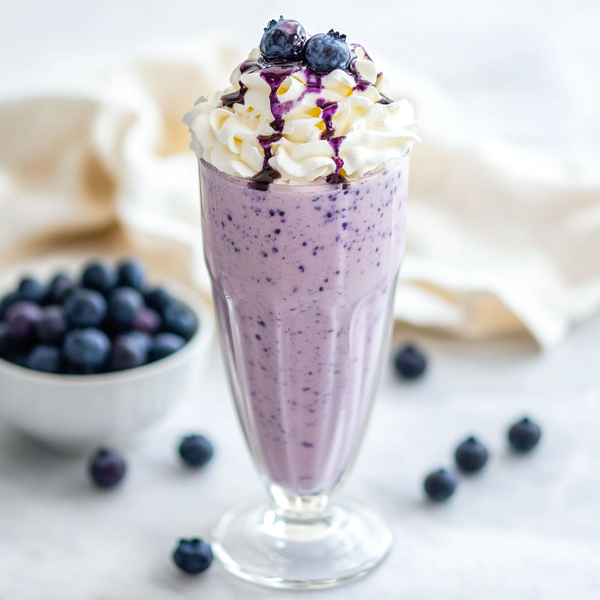 A slightly angled shot of a tall glass filled with a creamy blueberry vanilla milkshake, topped with fluffy whipped cream, a drizzle of blueberry syrup, and fresh blueberries. The glass sits on a white marble surface with a soft beige linen in the background, alongside a small bowl of fresh blueberries. The lighting is bright and natural, highlighting the smooth texture of the milkshake and vibrant blue tones.