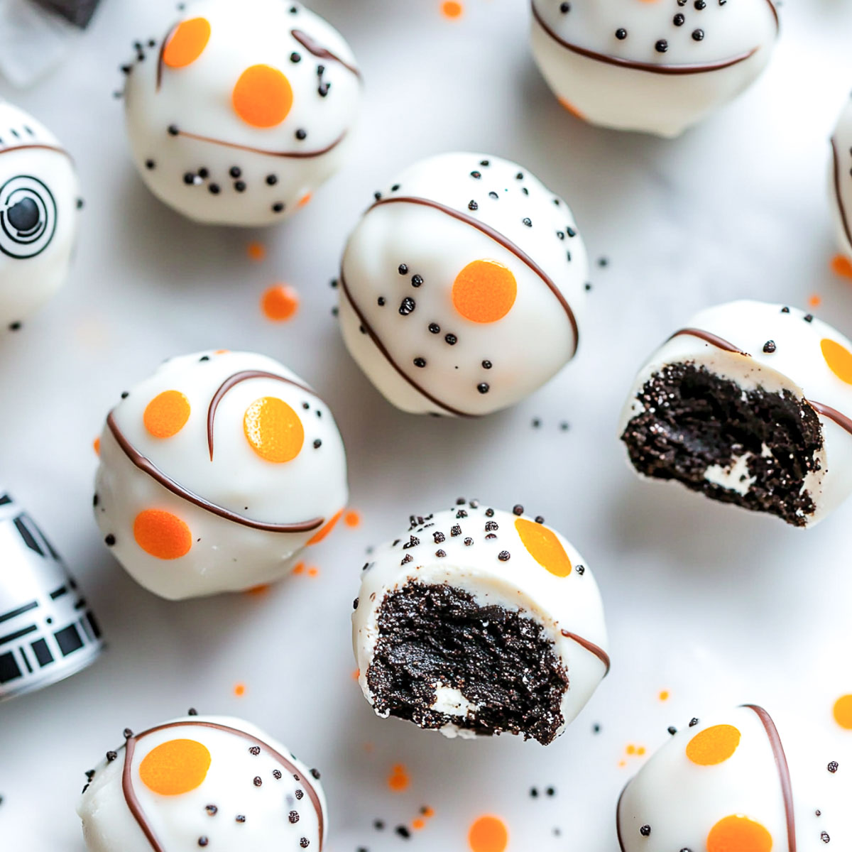A top-down shot of BB-8 themed Oreo truffles, coated in smooth white chocolate with orange and black details, arranged on a white marble surface. Some truffles are whole, while a few are cut in half, revealing a rich Oreo cream cheese filling. Small chocolate sprinkles and drizzles of orange candy melt enhance the BB-8 droid design.