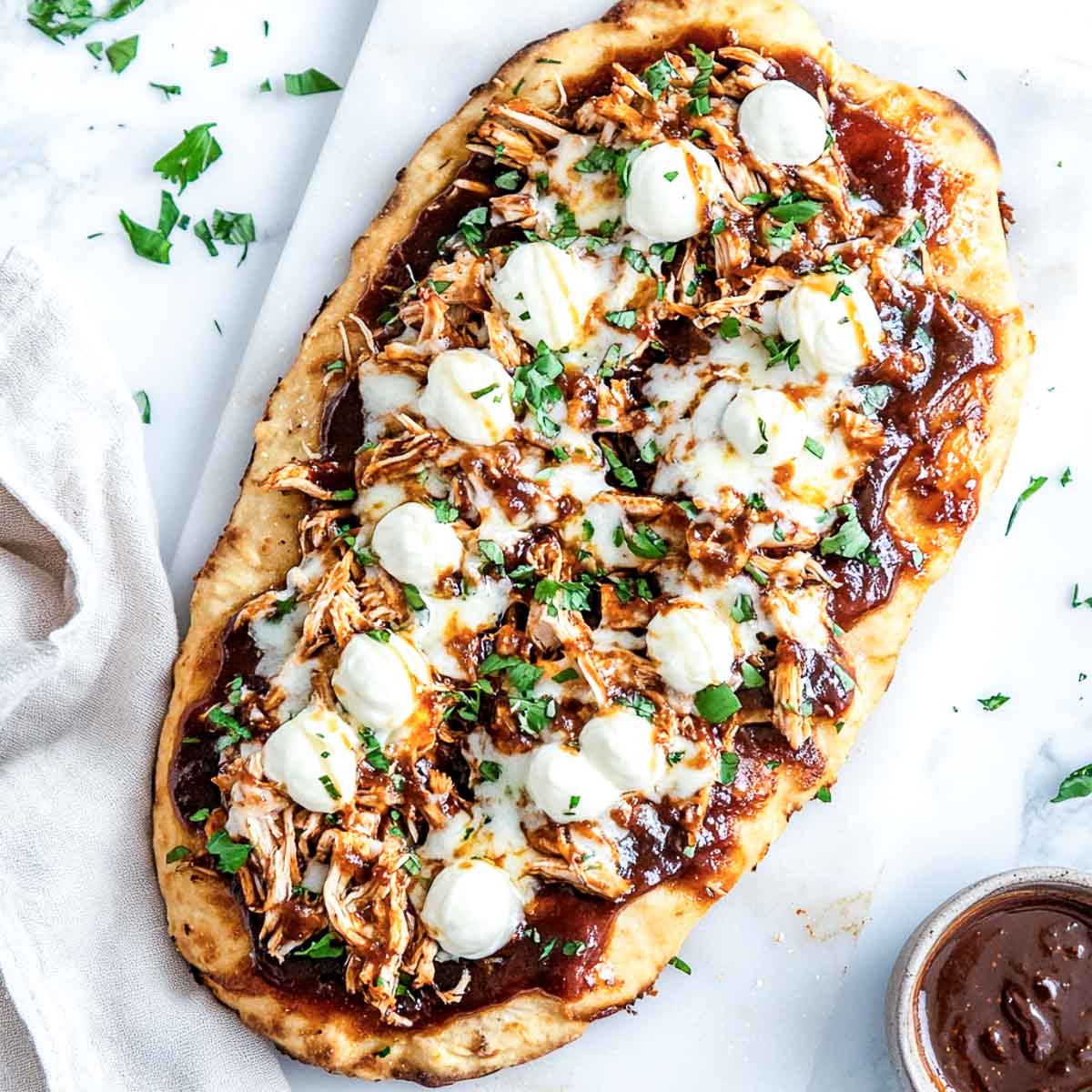 A top-down image of a rustic, golden-brown flatbread topped with BBQ sauce, shredded chicken, and melted mozzarella pearls, arranged in a diagonal bandolier pattern. The flatbread is placed on parchment paper over a white marble surface, with a small bowl of extra BBQ sauce and scattered fresh parsley leaves beside it.