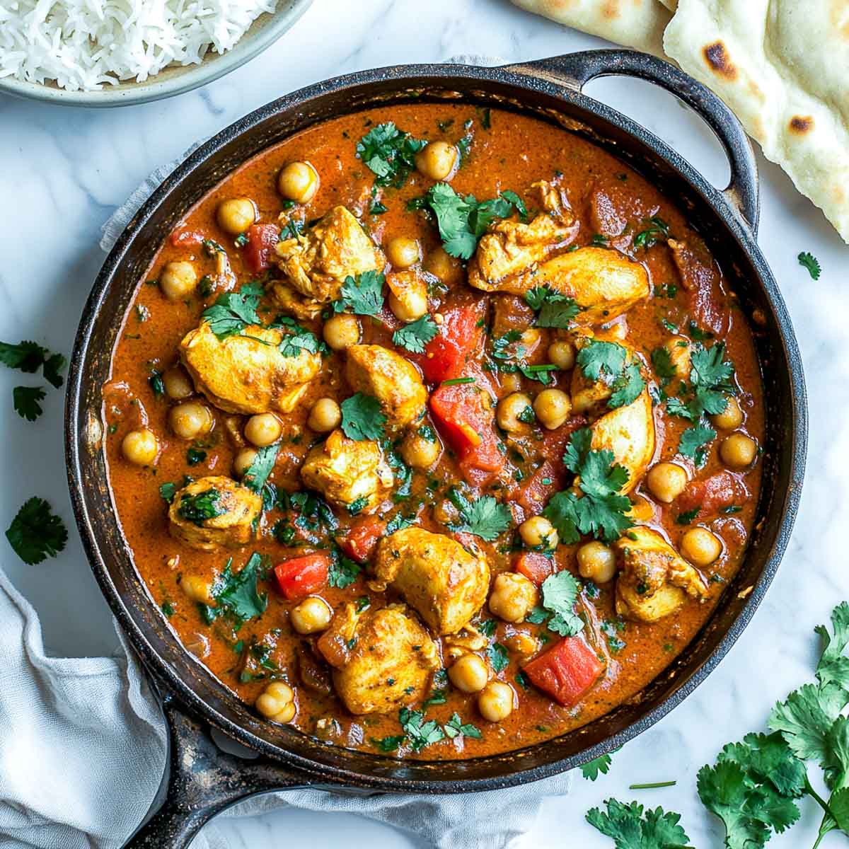 Chicken chickpea curry in a skillet with rice and naan in the background.