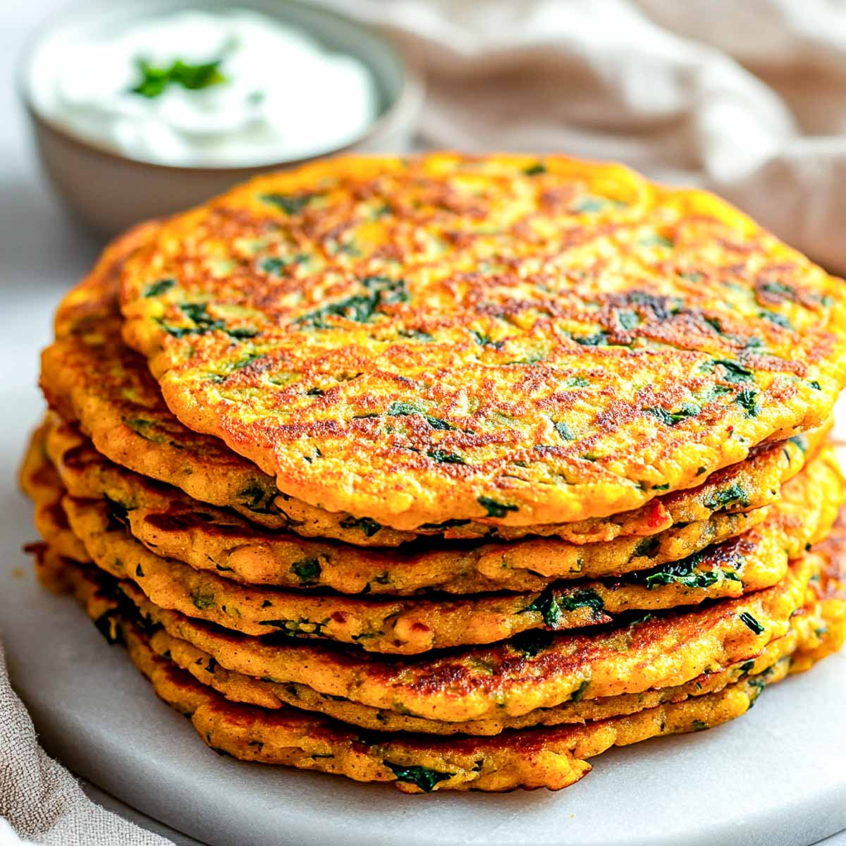 A stack of golden savory chickpea pancakes with spinach, served on a white marble surface with a side of yogurt.