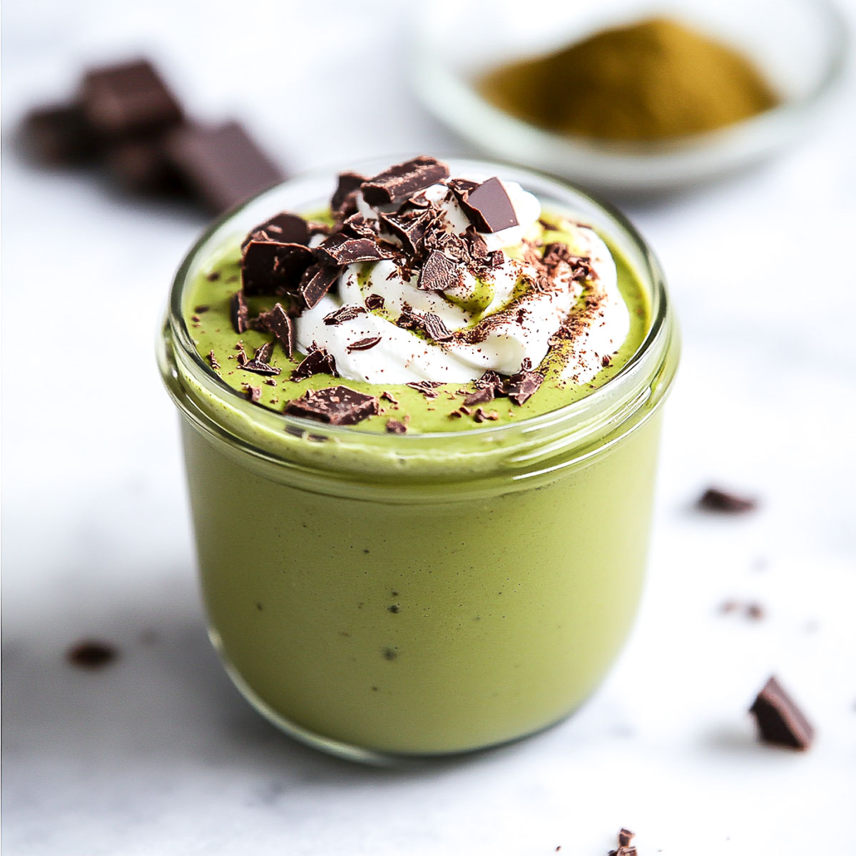A slightly angled side view of a matcha pudding served in a glass jar, topped with a swirl of whipped cream and dark chocolate shavings. The vibrant green color stands out against the marble background, with scattered chocolate pieces and a small bowl of matcha powder in the softly blurred background.