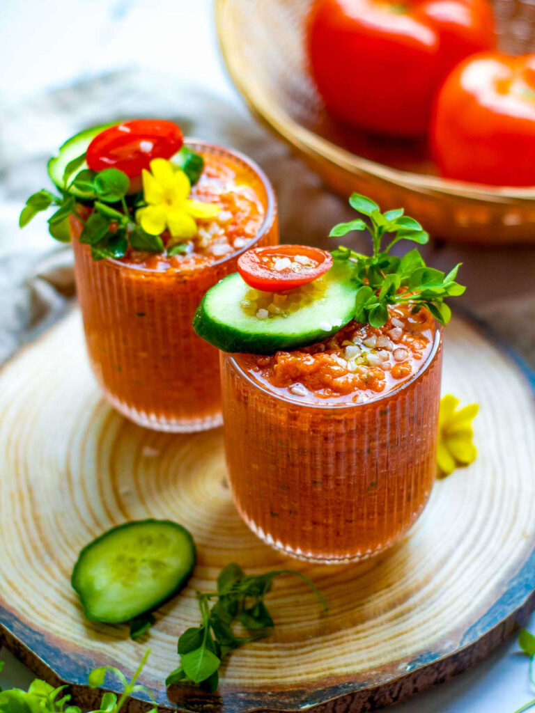 Two shot glasses of gazpacho placed on a wooden board with a basket of tomatoes in the background.
