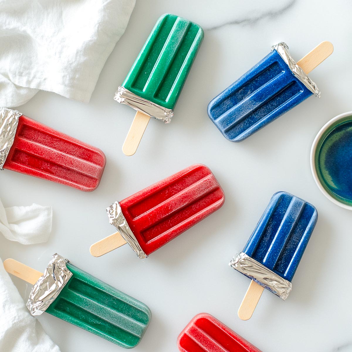A wide-angle image of glowing homemade popsicles in red, blue, and green, arranged on a white marble surface. The popsicles have foil-wrapped handles resembling lightsaber hilts. Their smooth, frosty texture is visible, with ice cubes scattered around. Bright, natural lighting enhances the vibrant neon colors.