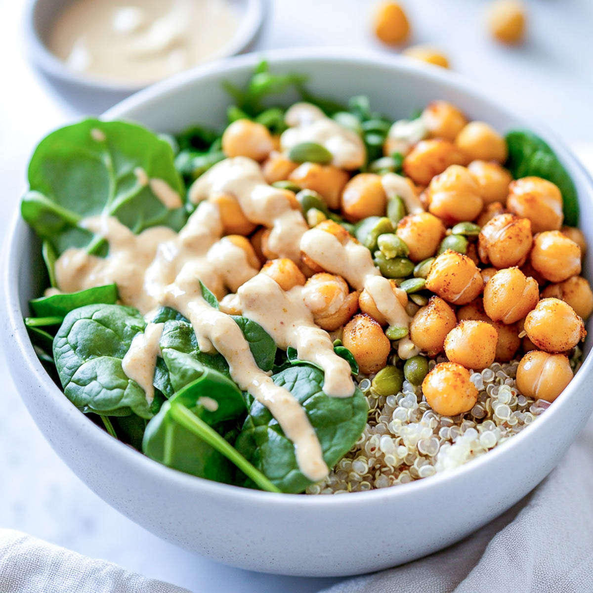A high-protein breakfast power bowl filled with quinoa, chickpeas, spinach, and tahini dressing, topped with pumpkin seeds, served in a white ceramic bowl on a white marble surface.