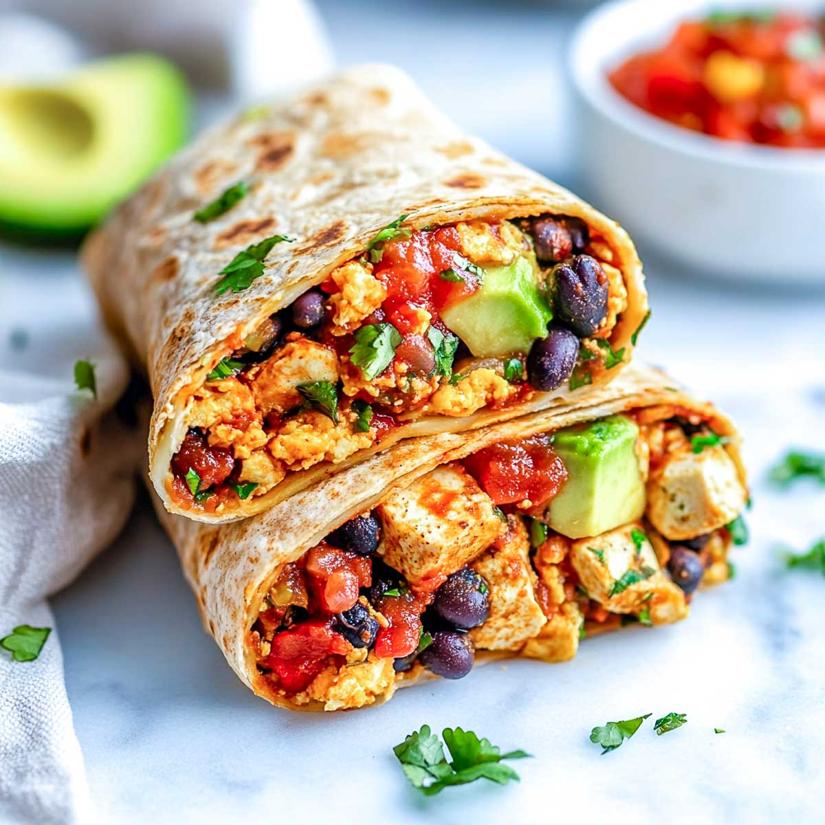 A whole wheat breakfast burrito sliced in half, filled with black beans, tofu, cheese, salsa, and avocado, placed on a white marble surface.