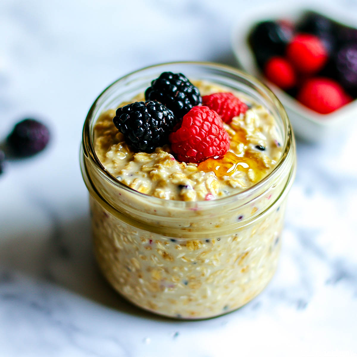 A glass jar filled with creamy overnight oats, topped with fresh berries and a drizzle of honey, placed on a white marble surface with a spoon resting beside it.
