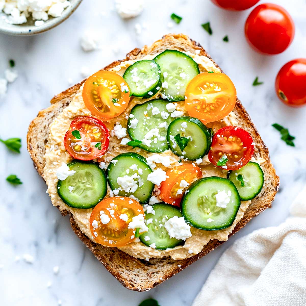 A slice of whole grain toast topped with hummus, cucumber slices, cherry tomatoes, and crumbled feta, placed on a white marble surface.