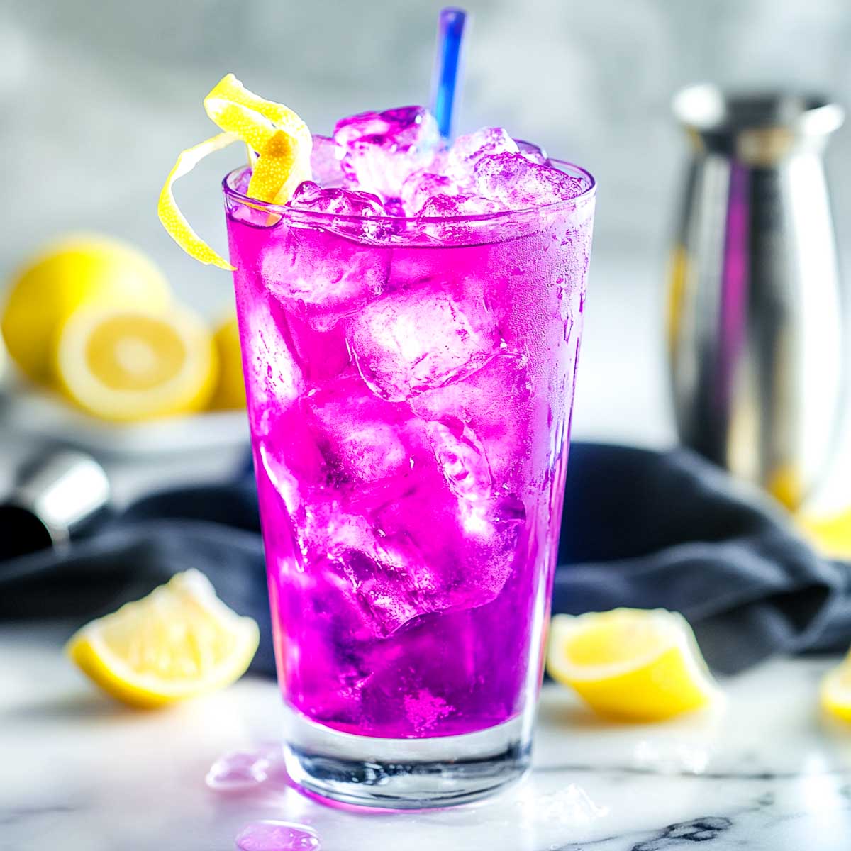 A slightly angled side view of a bright neon-purple cocktail served in a highball glass on a white marble surface. The drink is filled with crushed ice, giving it an electrifying glow. A twisted lemon peel rests on the rim, and a blue swizzle stick adds a futuristic touch. In the softly blurred background, fresh lemons, a cocktail jigger, and a black linen napkin enhance the cosmic, sci-fi aesthetic.