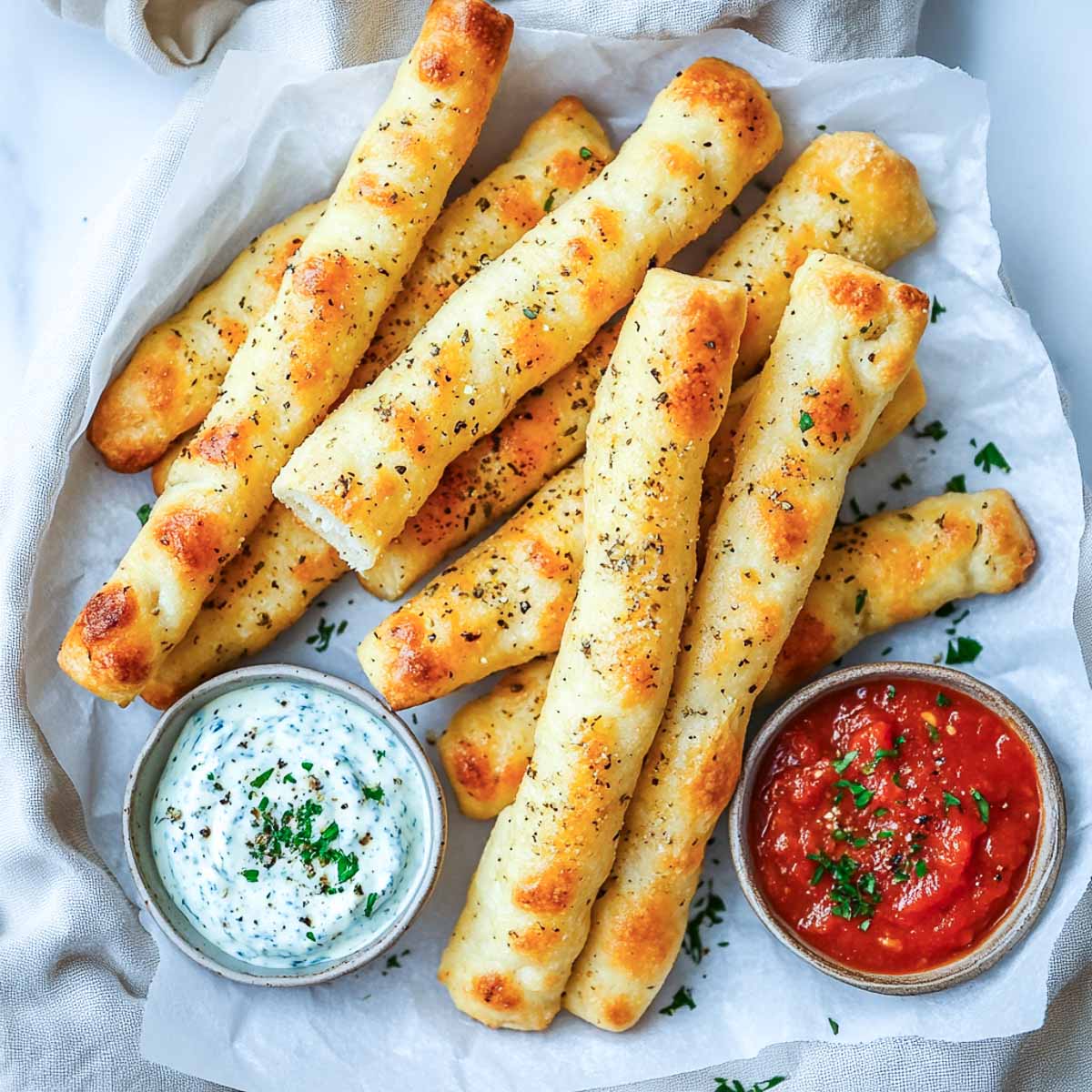 A top-down image of golden, crispy breadsticks sprinkled with herbs, served with two Star Wars-themed dipping sauces—one deep red Dark Side Marinara and one creamy Blue Holocron Dip—in small ceramic bowls on a white marble background.