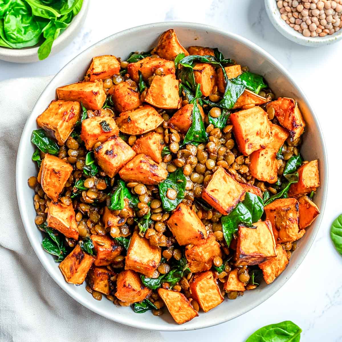 A hearty lentil and sweet potato hash with sautéed spinach, served in a white ceramic bowl on a white marble surface.