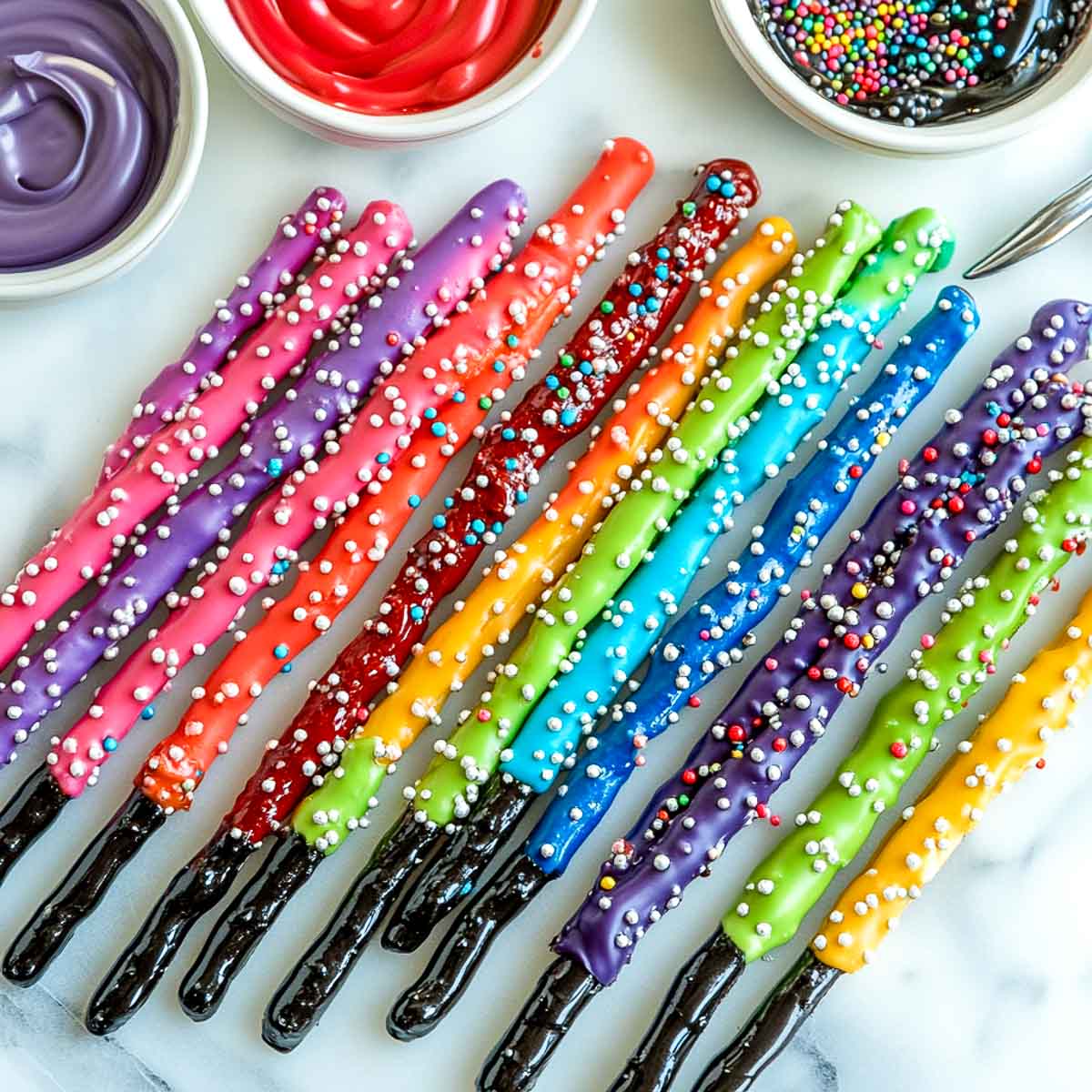 A row of candy-coated pretzel rods resembling lightsabers, dipped in vibrant red, blue, green, and purple coatings. The bottom portion of each pretzel is coated in dark chocolate to create the handle. The pretzels are arranged on a white marble surface, with a few scattered sprinkles nearby.