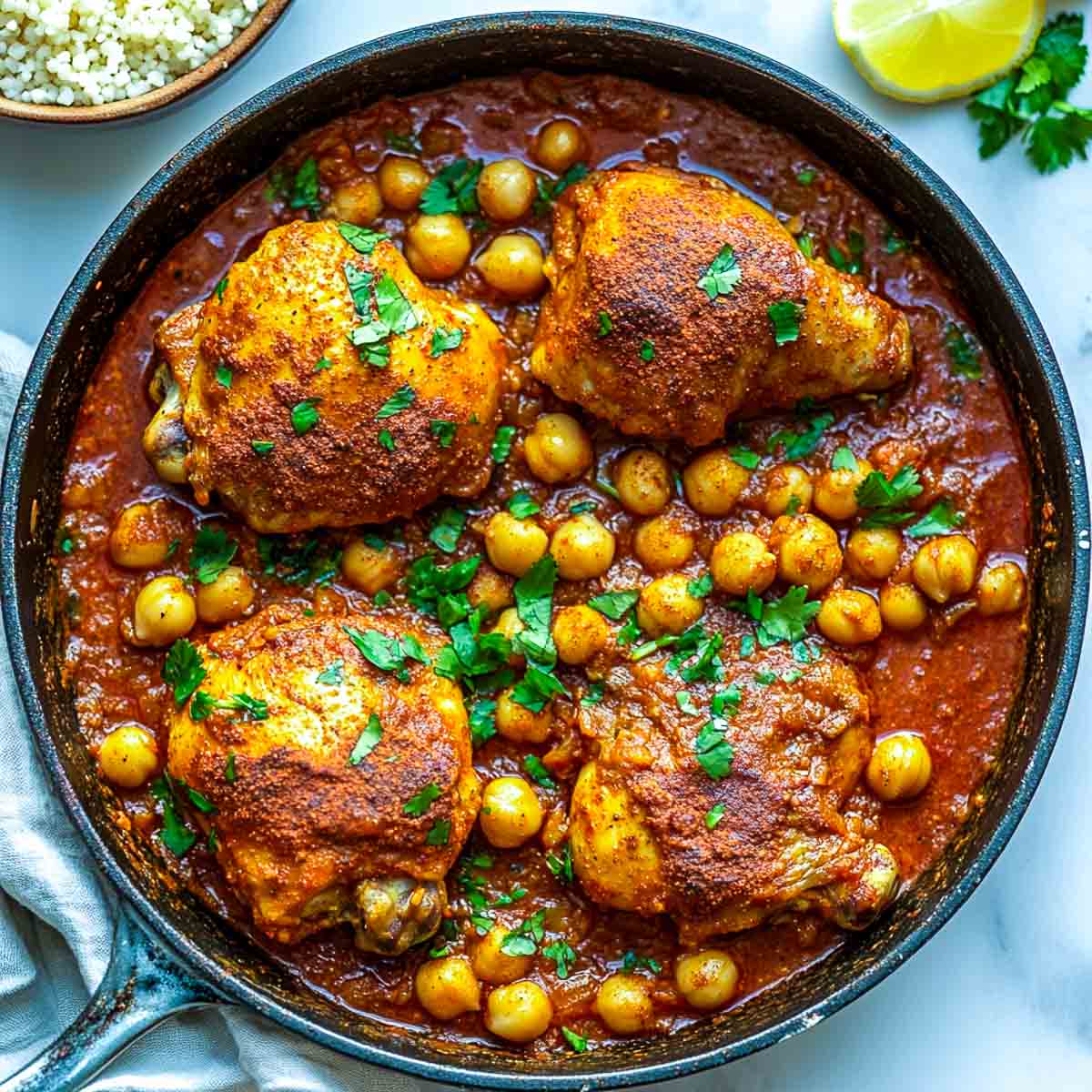 Moroccan chicken and chickpeas in a skillet with a bowl of rice in the background.