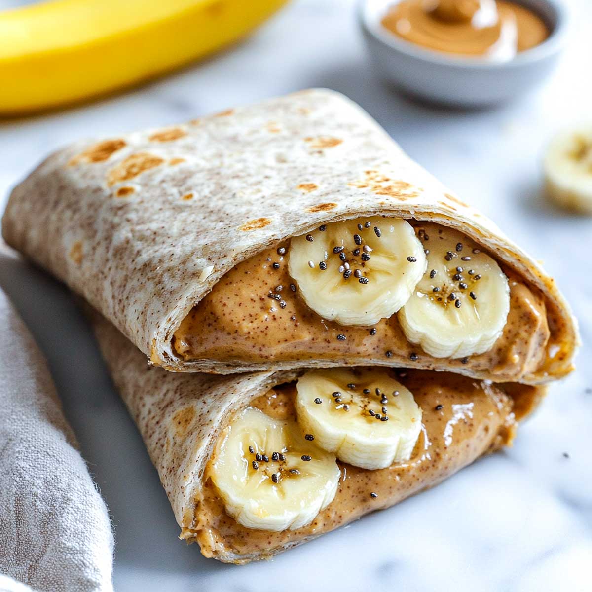 A peanut butter banana protein wrap sliced in half, showing creamy peanut butter, banana slices, chia seeds, and hemp seeds inside a whole wheat tortilla, placed on a white marble surface.
