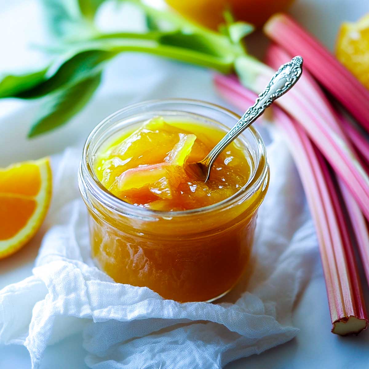 A small glass jar of rhubarb orange jam with a spoon with rhubard in the background.