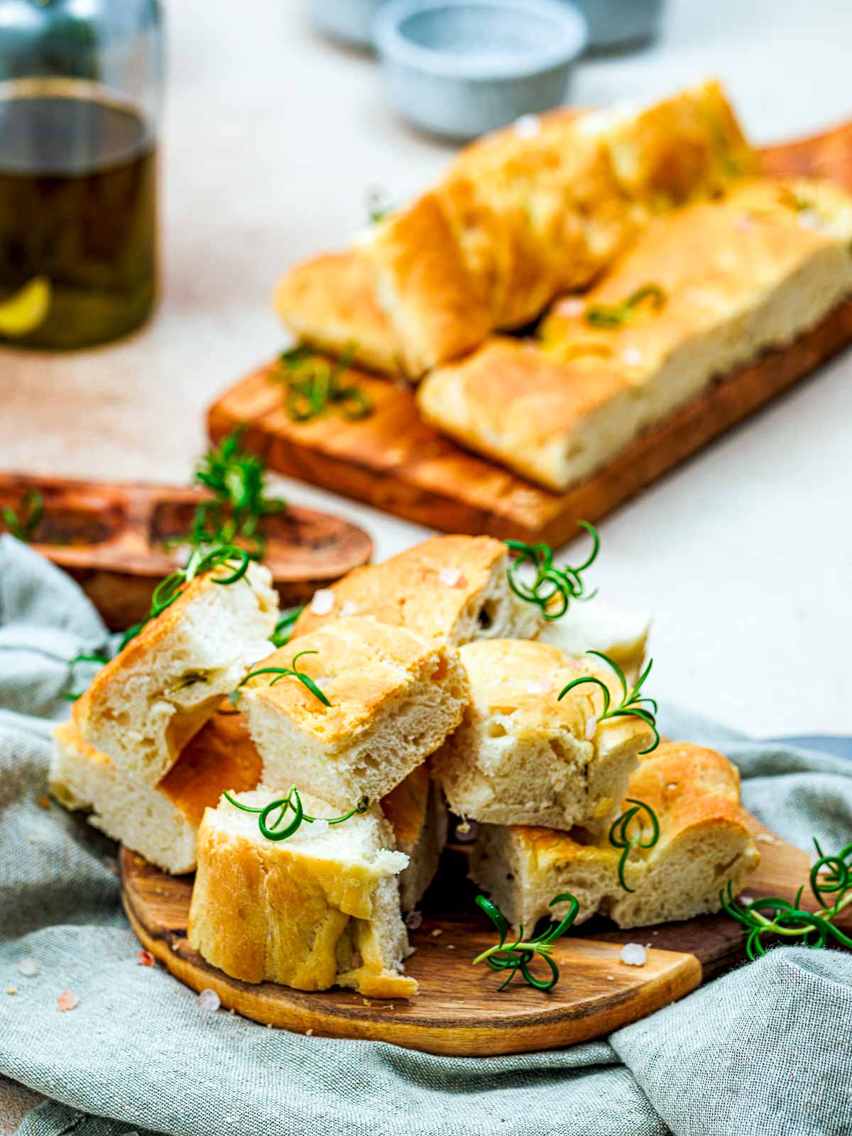 Small square pieces of rosemary focaccia on a wooden board with remaning focaccia and olive oil in the background.