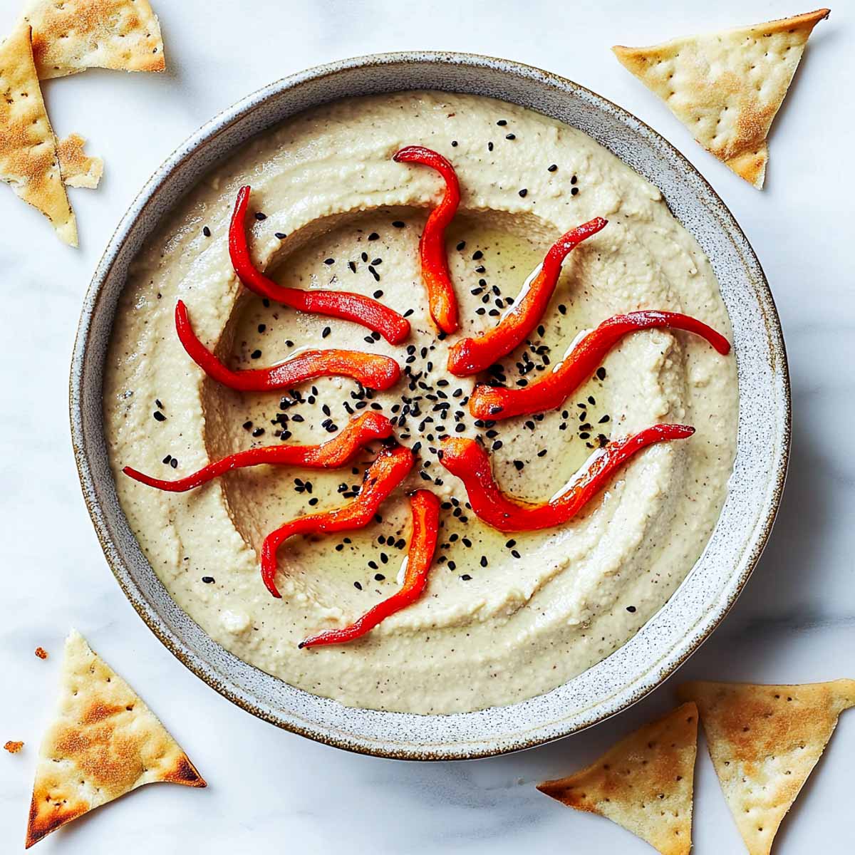 A shallow dish of creamy hummus designed to resemble a pit in the desert, with pita chips arranged around the edges. Thin strips of roasted red pepper curl out from the center like tentacles, and a sprinkle of black sesame seeds adds texture. The dish sits on a white marble surface, with a few extra pita chips scattered nearby.
