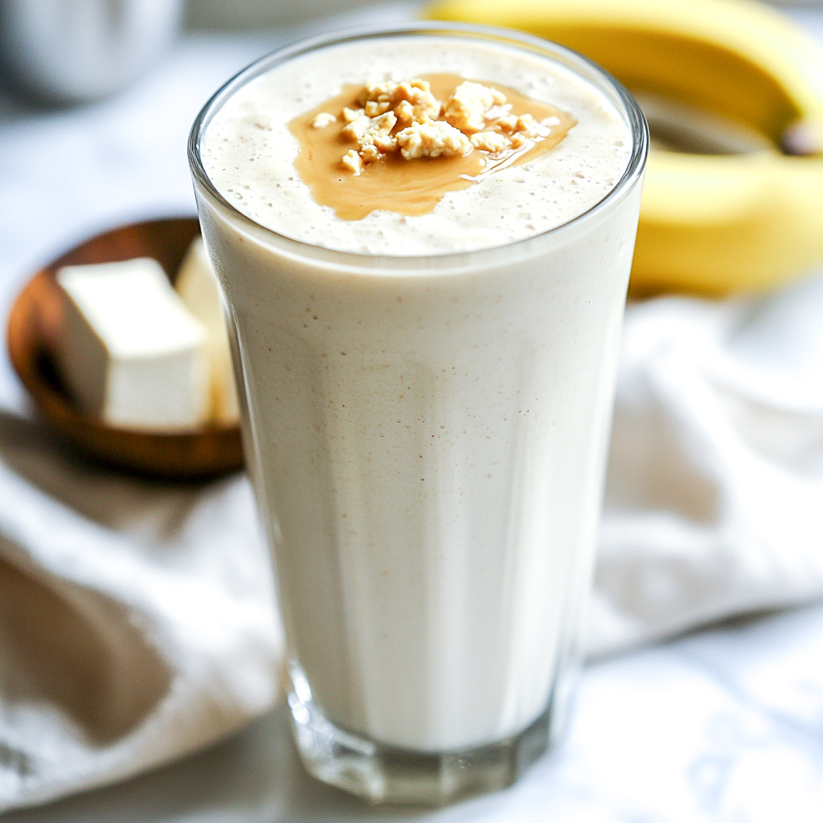 A tall glass filled with a creamy silken tofu protein shake, placed on a white marble surface with a drizzle of almond butter on top.