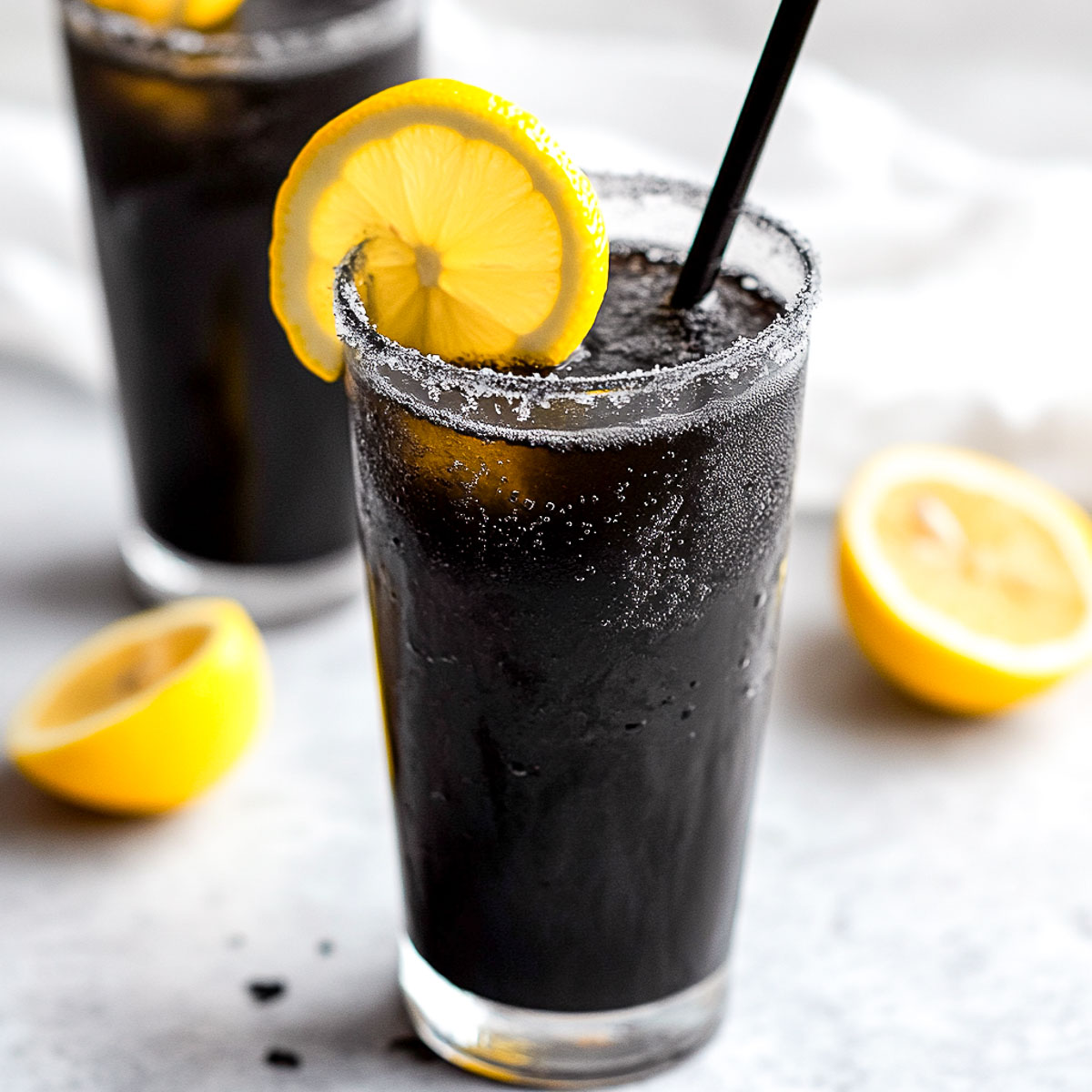 A slightly angled image of a tall frosted glass filled with dark black lemonade, rimmed with sugar, and garnished with a bright yellow lemon slice. Another glass is slightly blurred in the background, along with fresh lemons and a soft beige linen napkin. The surface is a cool-toned white marble, creating a sleek contrast with the deep black drink.