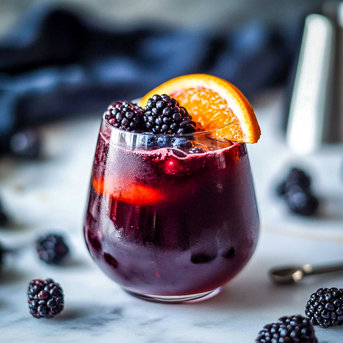 A deep red sangria served in a stemless wine glass on a white marble surface. The drink is filled with fresh blackberries, blueberries, and a thin orange slice, floating gently on top. A small black linen napkin, a metal cocktail jigger, and scattered dark berries complete the moody, Sith-like aesthetic.