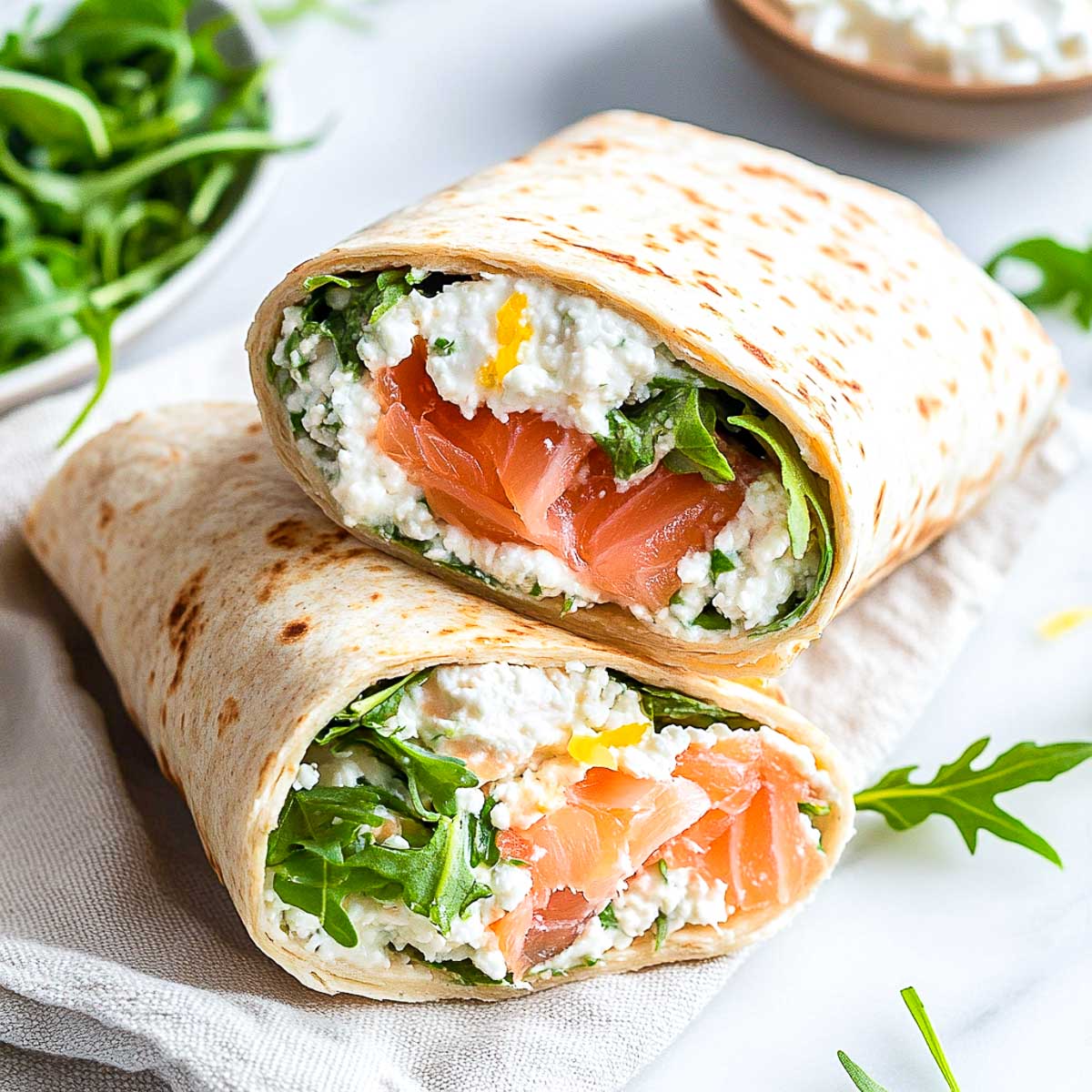 A whole wheat wrap sliced in half, filled with smoked salmon, cottage cheese, and arugula, placed on a white marble surface.