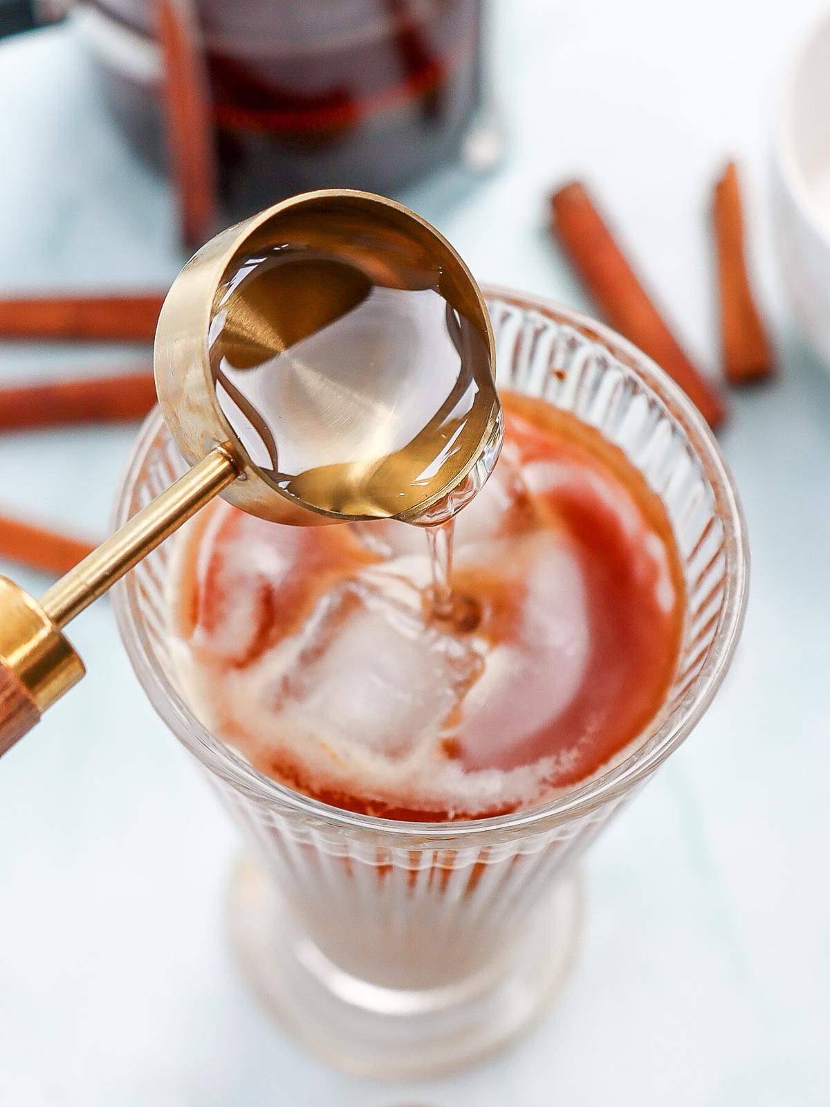 Golden measuring spoon pouring homemade cinnamon dolce syrup into a glass of iced coffee with ice cubes, cinnamon sticks, and a French press in the background.