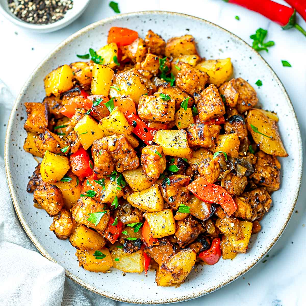 A plate of tempeh breakfast hash with crispy potatoes and sautéed bell peppers, served on a white marble surface.