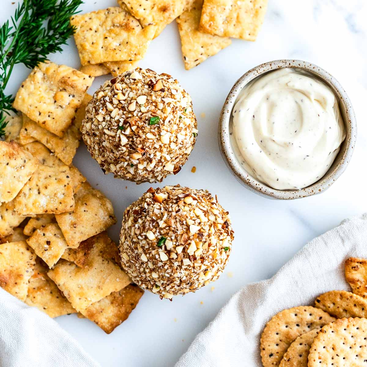A wide-angle top-down shot of two golden cheese balls coated in chopped nuts and fresh herbs, placed on a white marble surface. A bowl of creamy dip sits beside them, surrounded by an assortment of crackers, creating a rustic, homemade party spread.