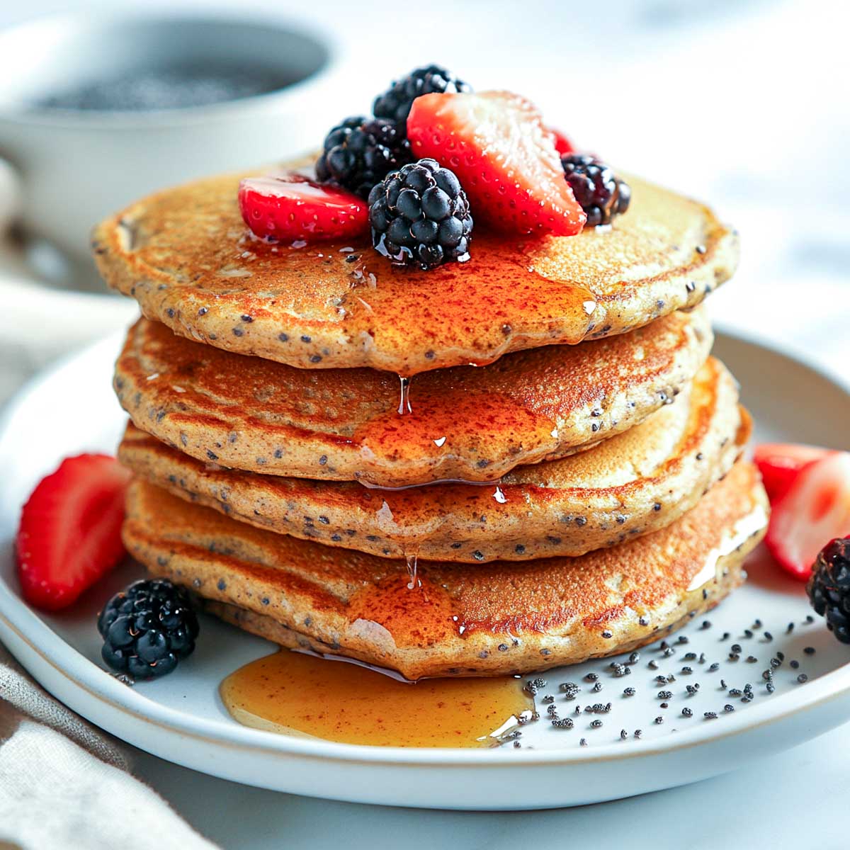 A stack of fluffy vegan protein pancakes on a white ceramic plate, topped with maple syrup, fresh berries, and a sprinkle of chia seeds, placed on a white marble surface.
