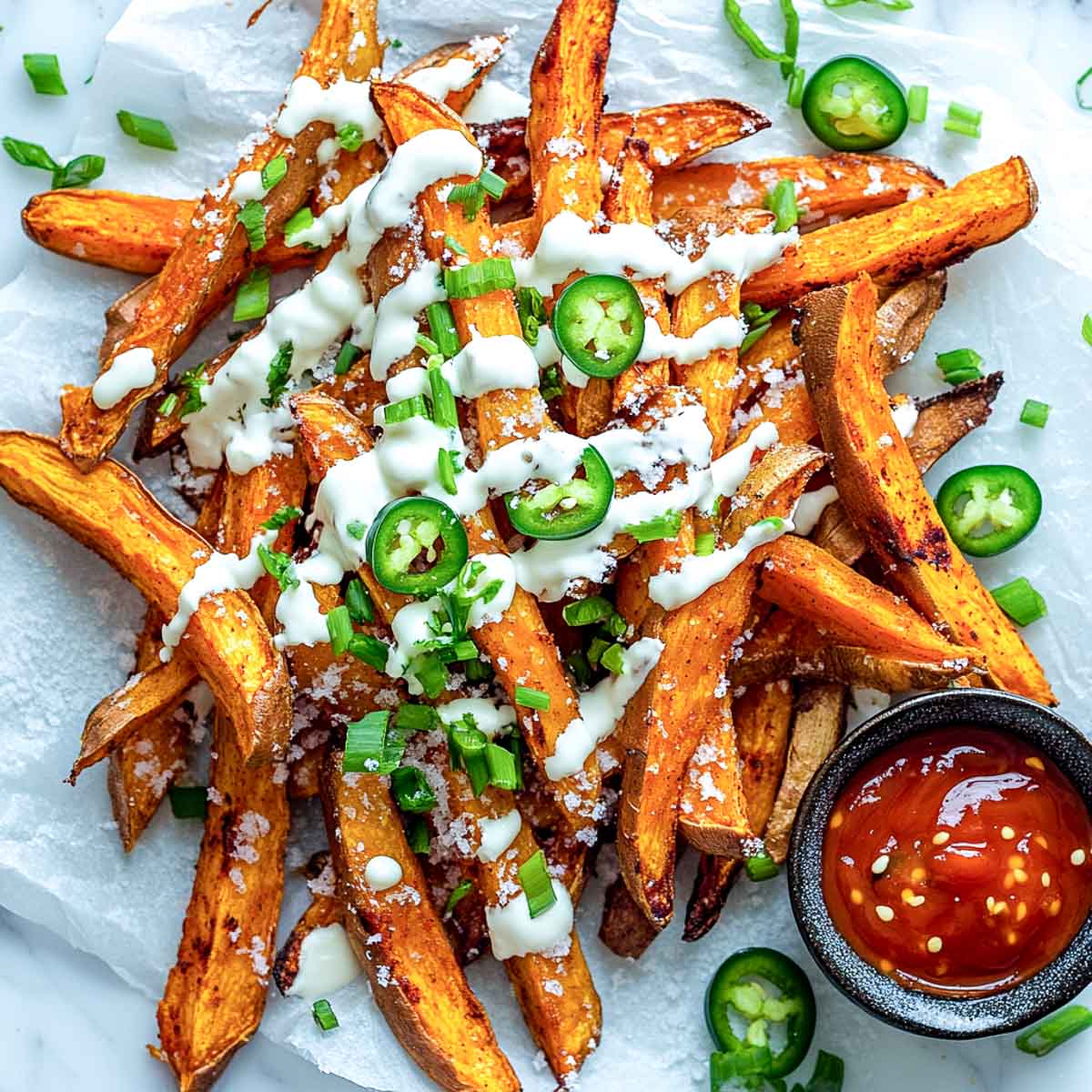 A top-down image of crispy, golden sweet potato fries piled on a white marble surface, drizzled with sour cream to resemble snow, topped with melted cheese, sliced jalapeños, and green onions. A small bowl of fiery red dipping sauce sits nearby, representing the Wampa’s fury. The scene has a slightly messy, homemade feel, with a napkin and scattered ingredients adding depth. Bright, natural lighting enhances the textures and colors.