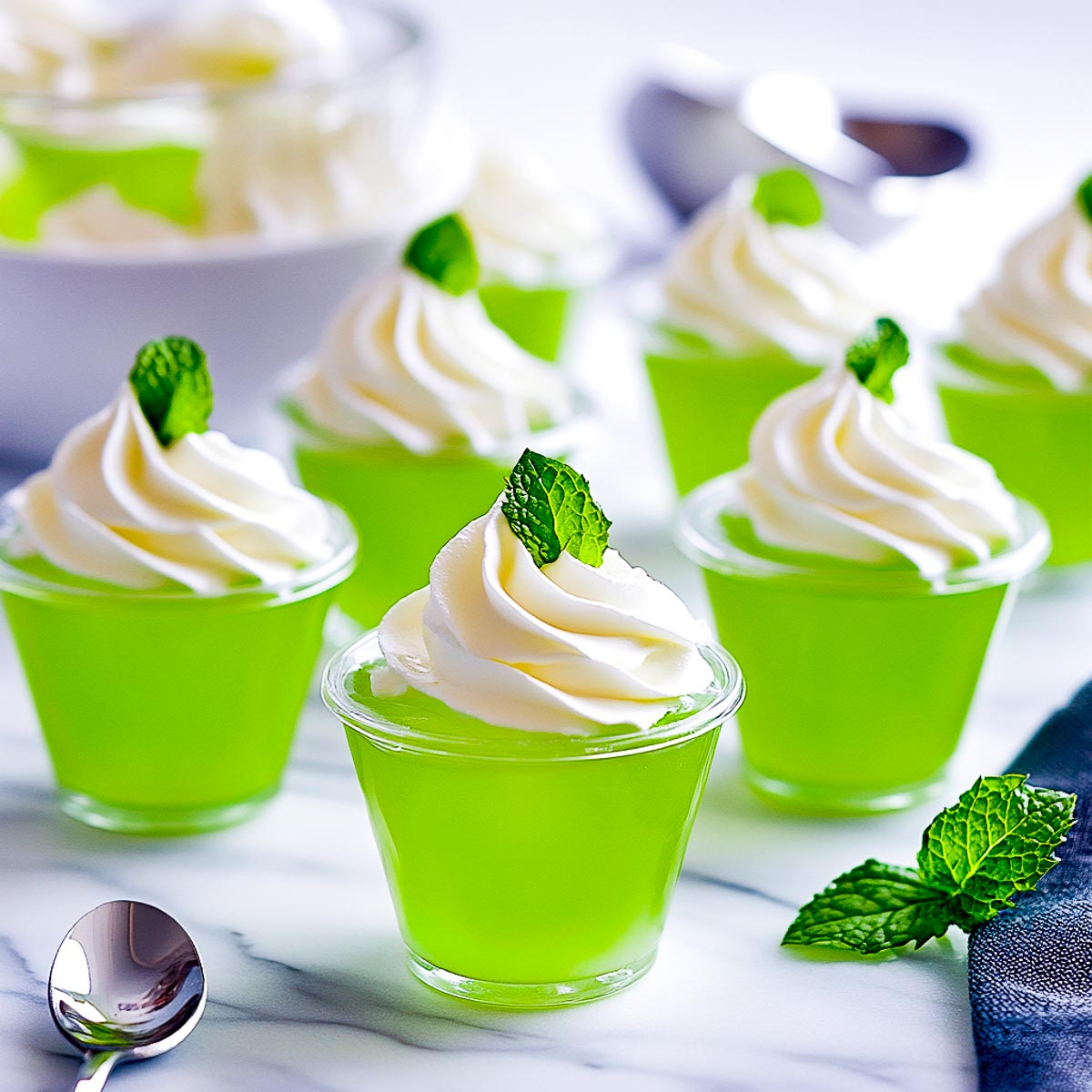 A wide-angle image of small clear cups filled with vibrant green Jello, topped with a soft swirl of Cool Whip and a fresh mint leaf garnish. The cups are arranged on a white marble surface, with a few scattered mint leaves and a spoon in the background, creating a clean and refreshing presentation.