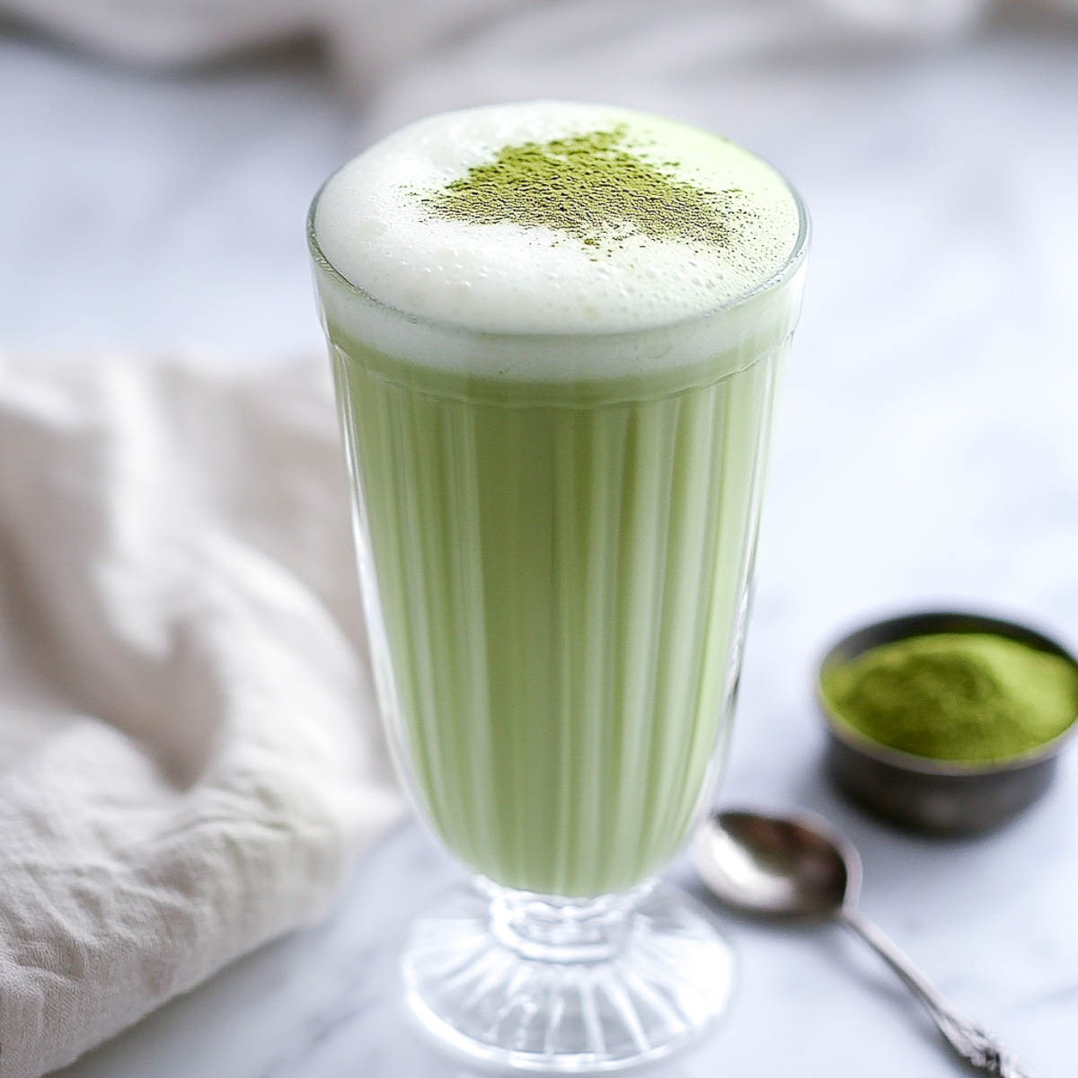 A slightly angled shot of a tall clear glass filled with a creamy, frothy green matcha latte, topped with a dusting of vibrant green matcha powder. The glass sits on a white marble surface, with a small rustic bowl of matcha powder in the background. A soft beige linen napkin drapes beside the glass, and a vintage-style spoon rests nearby, creating a warm and inviting scene.