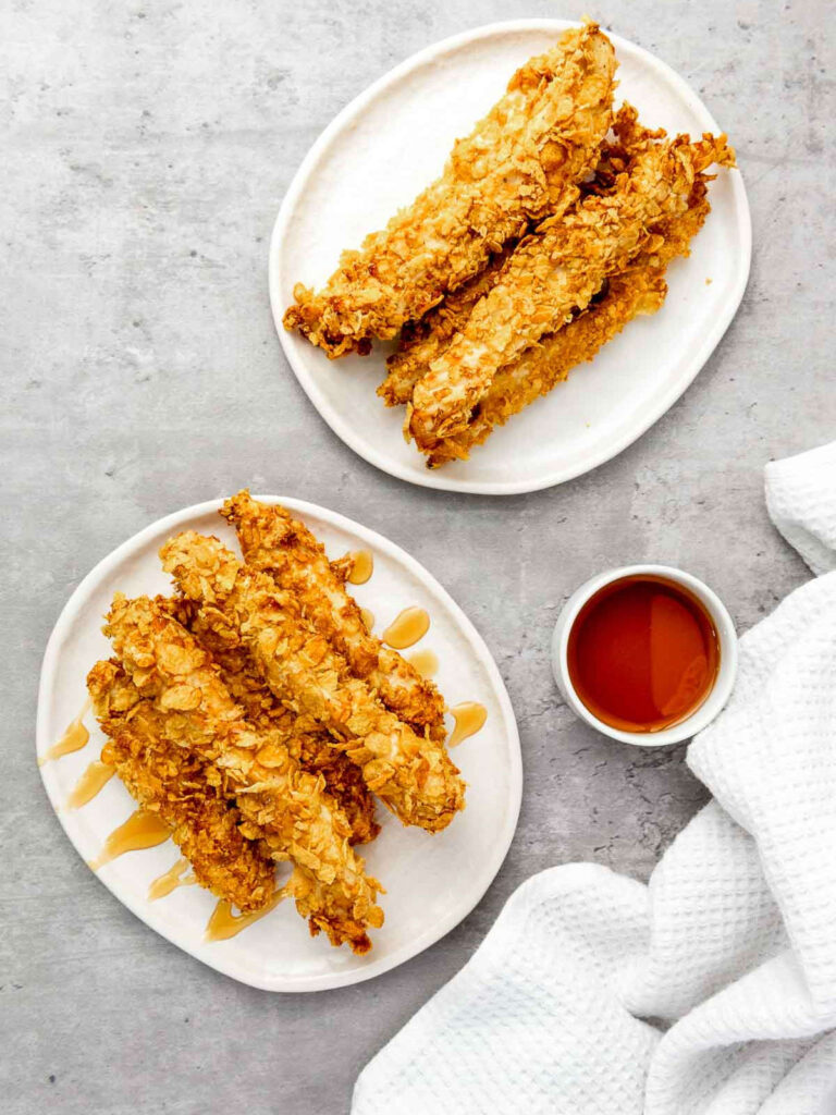 Two plates of crispy cornflake chicken served with hot honey on a grey surface.
