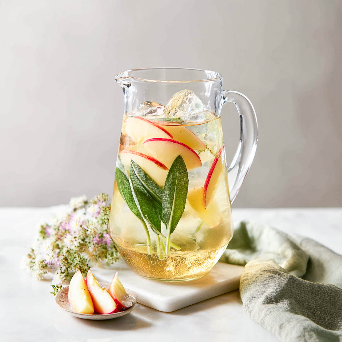 Pitcher of apple sage water with thin cored apple slices, sage leaves, and pale green napkin on marble surface.