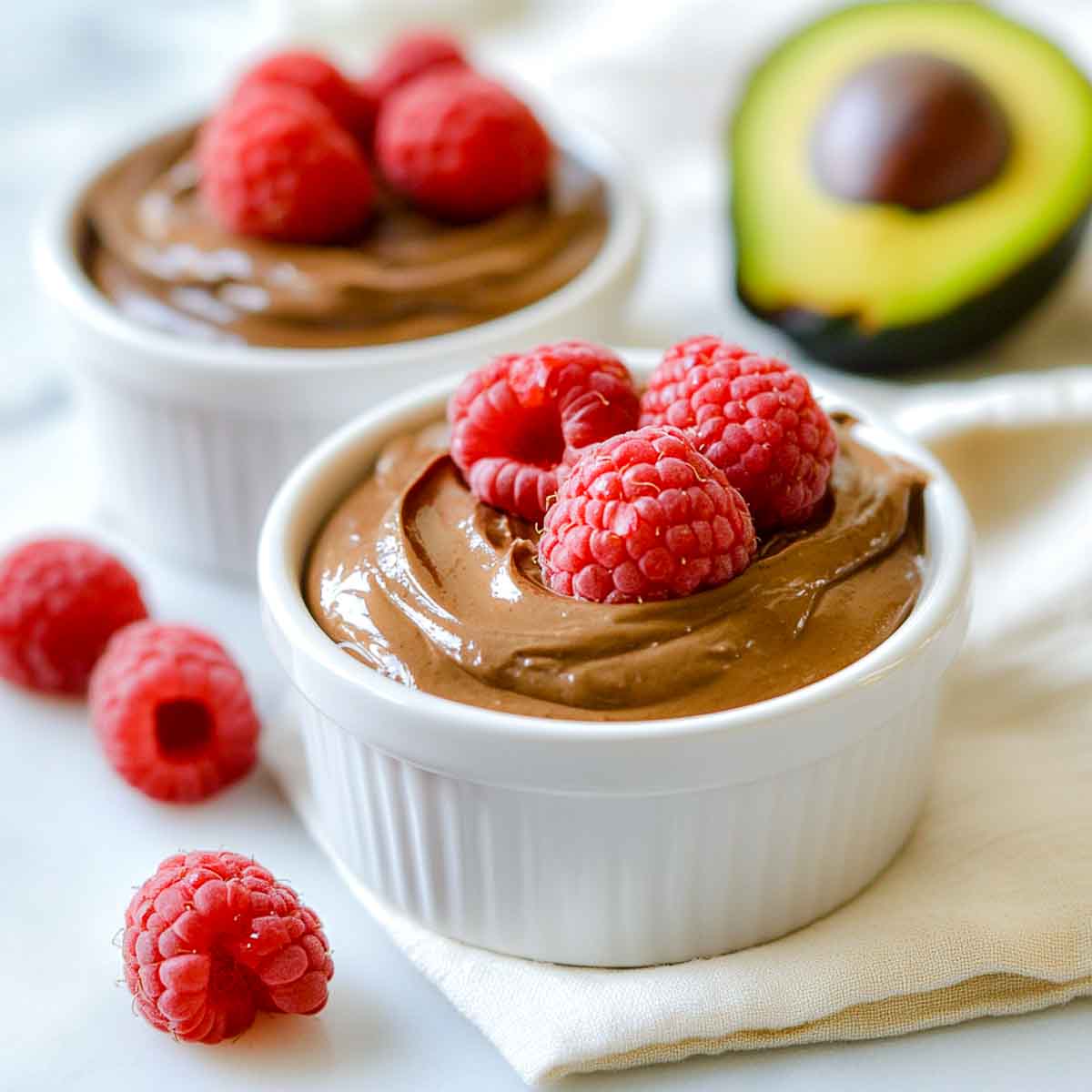 Two small white ramekins filled with silky chocolate avocado mousse topped with fresh raspberries.