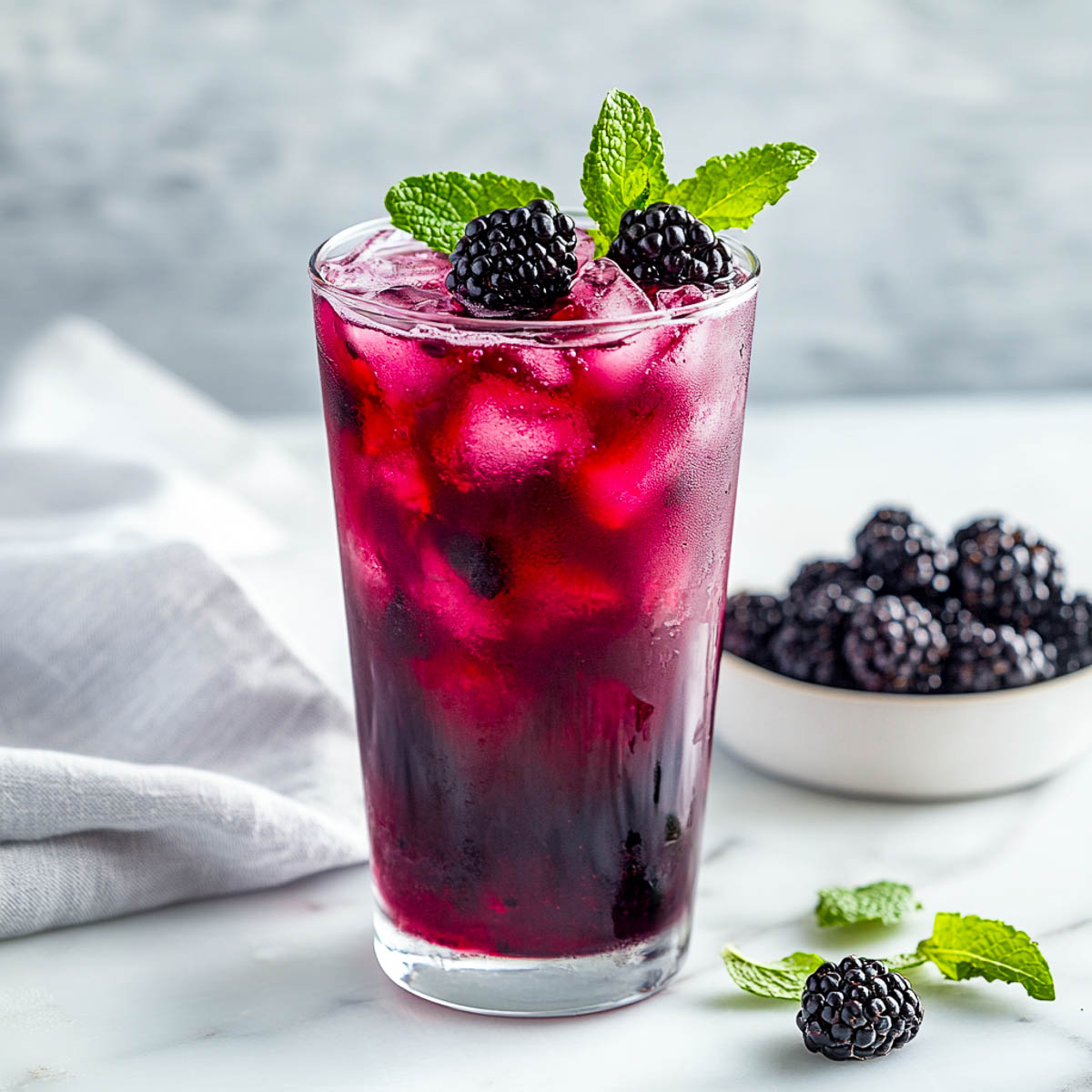 A tall glass of blackberry mint cooler with a bowl of blackberries in the background.