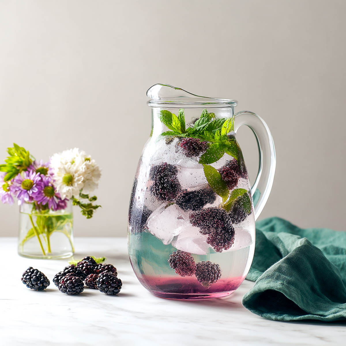 Pitcher of blackberry mint water with purple tint, mint leaves, dark berries, and green napkin beside a flower vase on marble surface.
