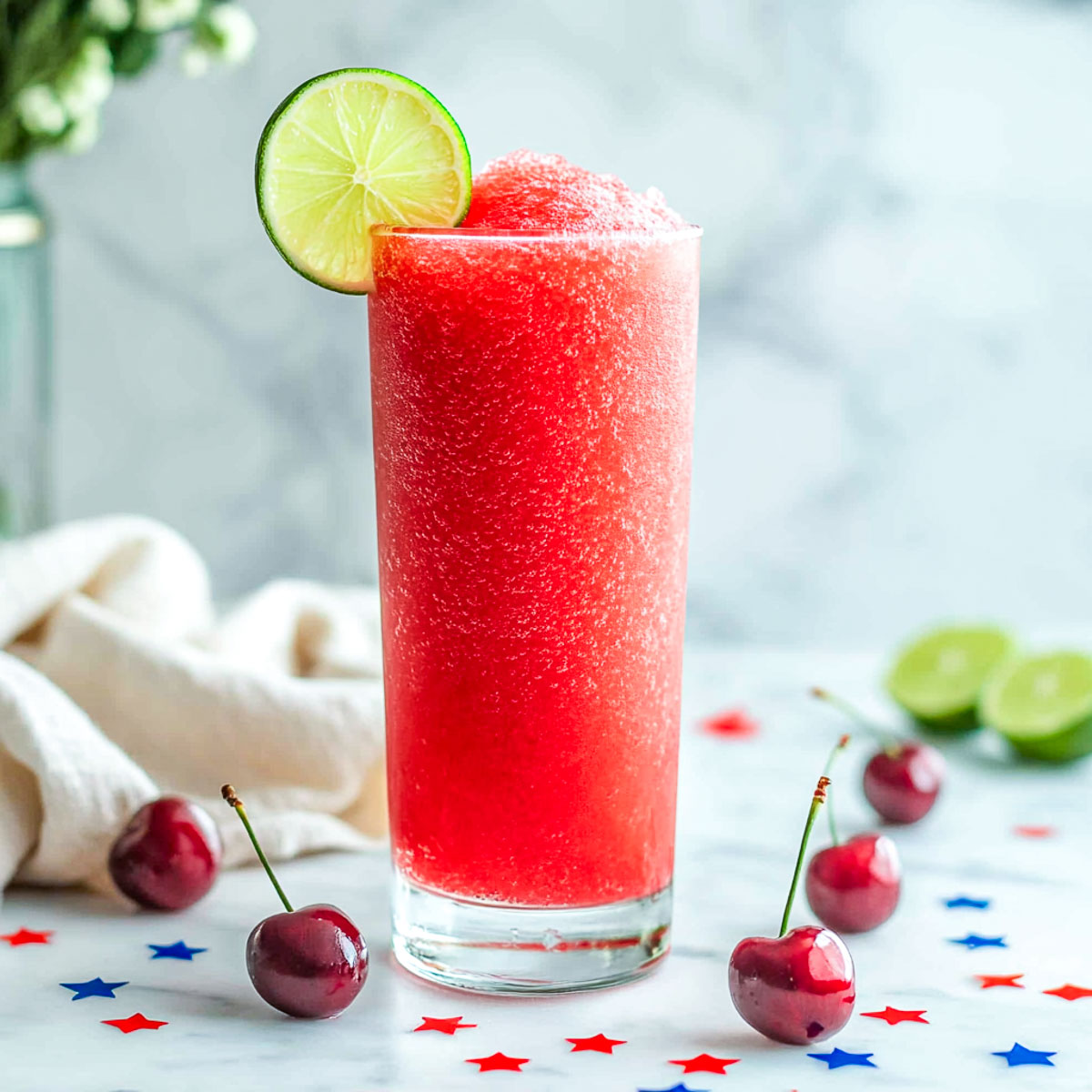 Tall glass of cherry limeade slush with lime slice and scattered cherries.