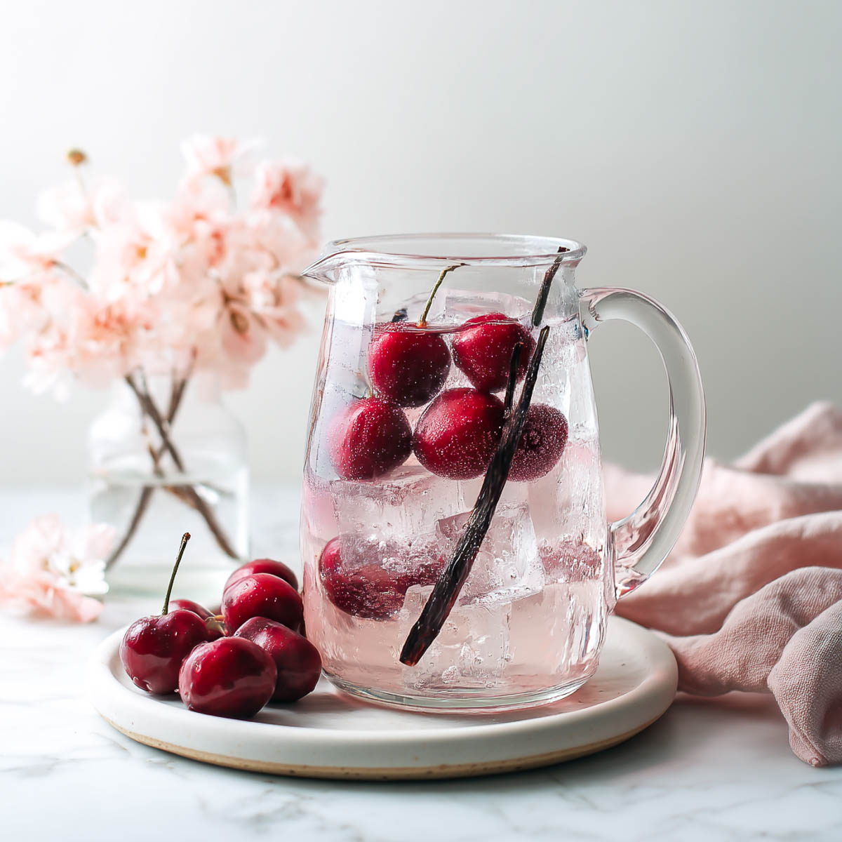 Pitcher of cherry vanilla water with dark cherries, split vanilla bean, blush napkin, and pink flowers on marble surface.